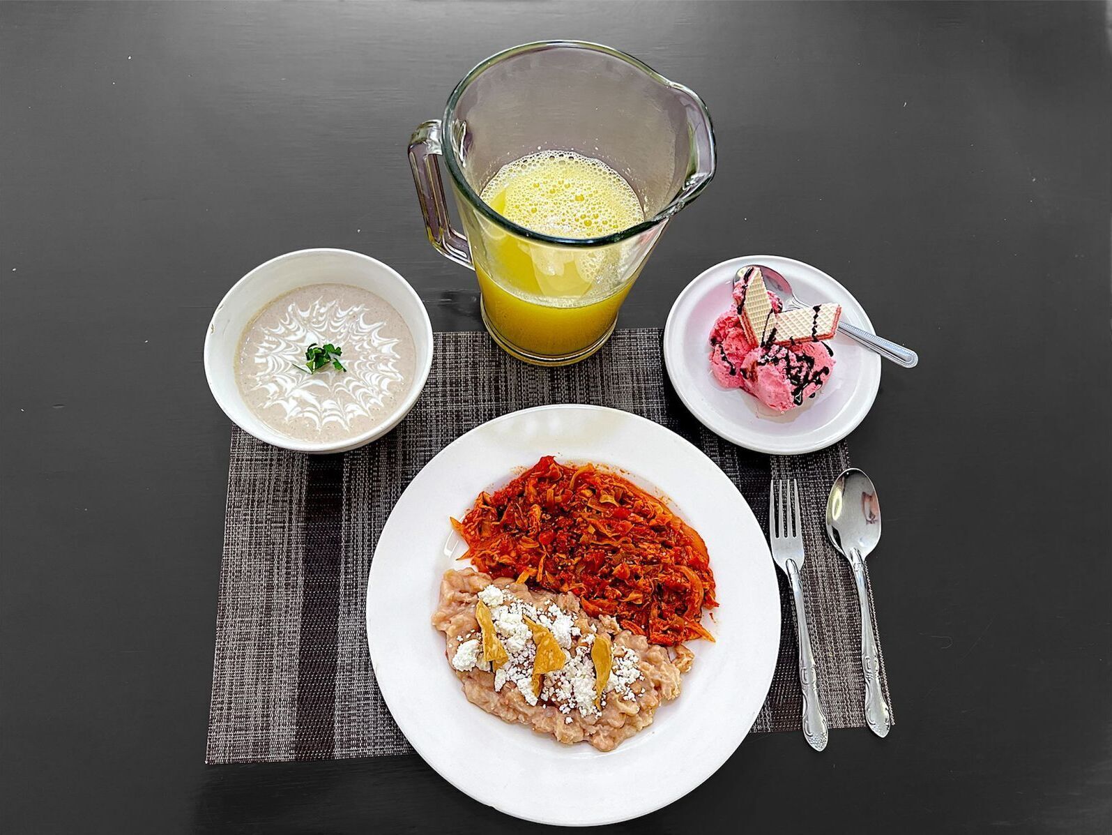 Une table dressée avec un repas composé d’un bol de soupe, d’une assiette de pâtes à la sauce tomate et au fromage, d’un verre de jus d’orange et d’une assiette de crème glacée.