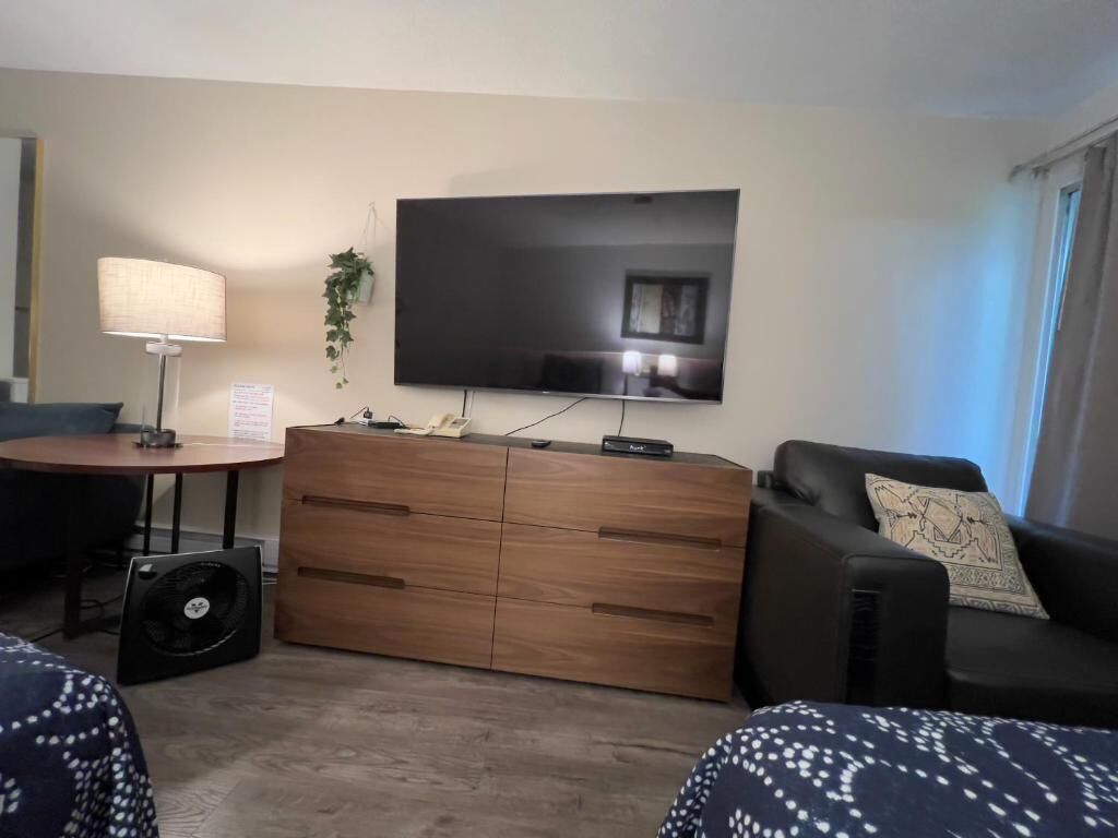A living room with a flat-screen TV, a wooden cabinet, a black sofa, and a patterned throw blanket.