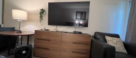 A living room with a flat-screen TV, a wooden cabinet, a black sofa, and a patterned throw blanket.