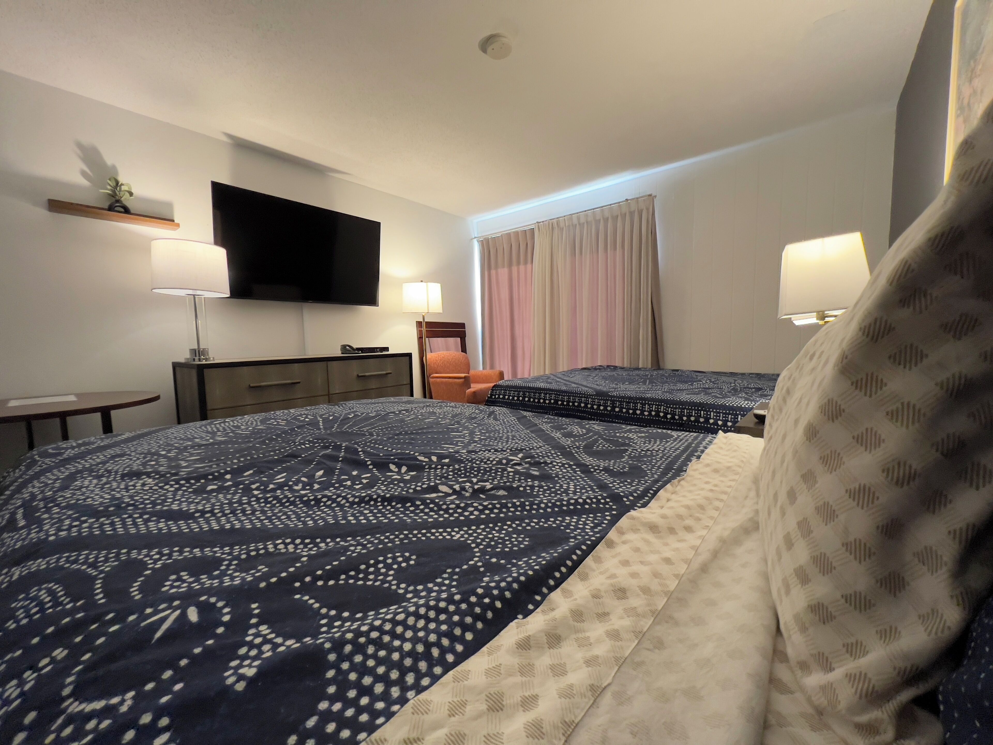 Habitación de hotel con una cama grande, televisión montada en la pared, mesita de noche, una silla y ventana con cortinas.