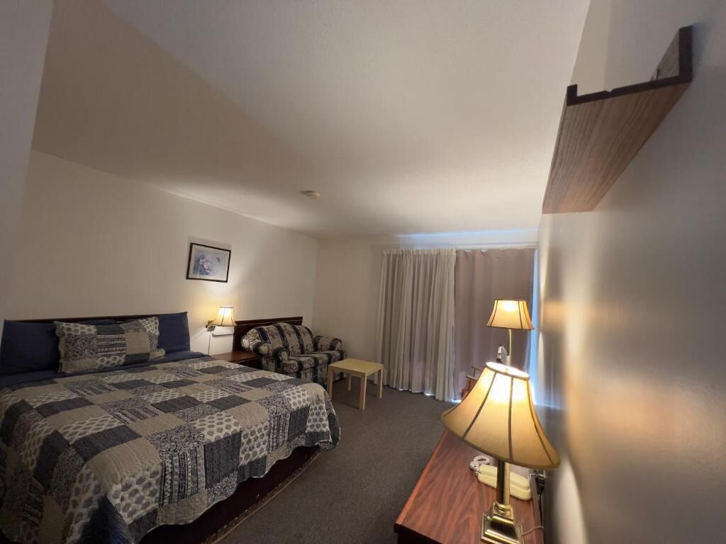 Habitación de hotel con cama, mesita de noche con lámpara, pequeña mesa, sofá y ventana con cortinas.