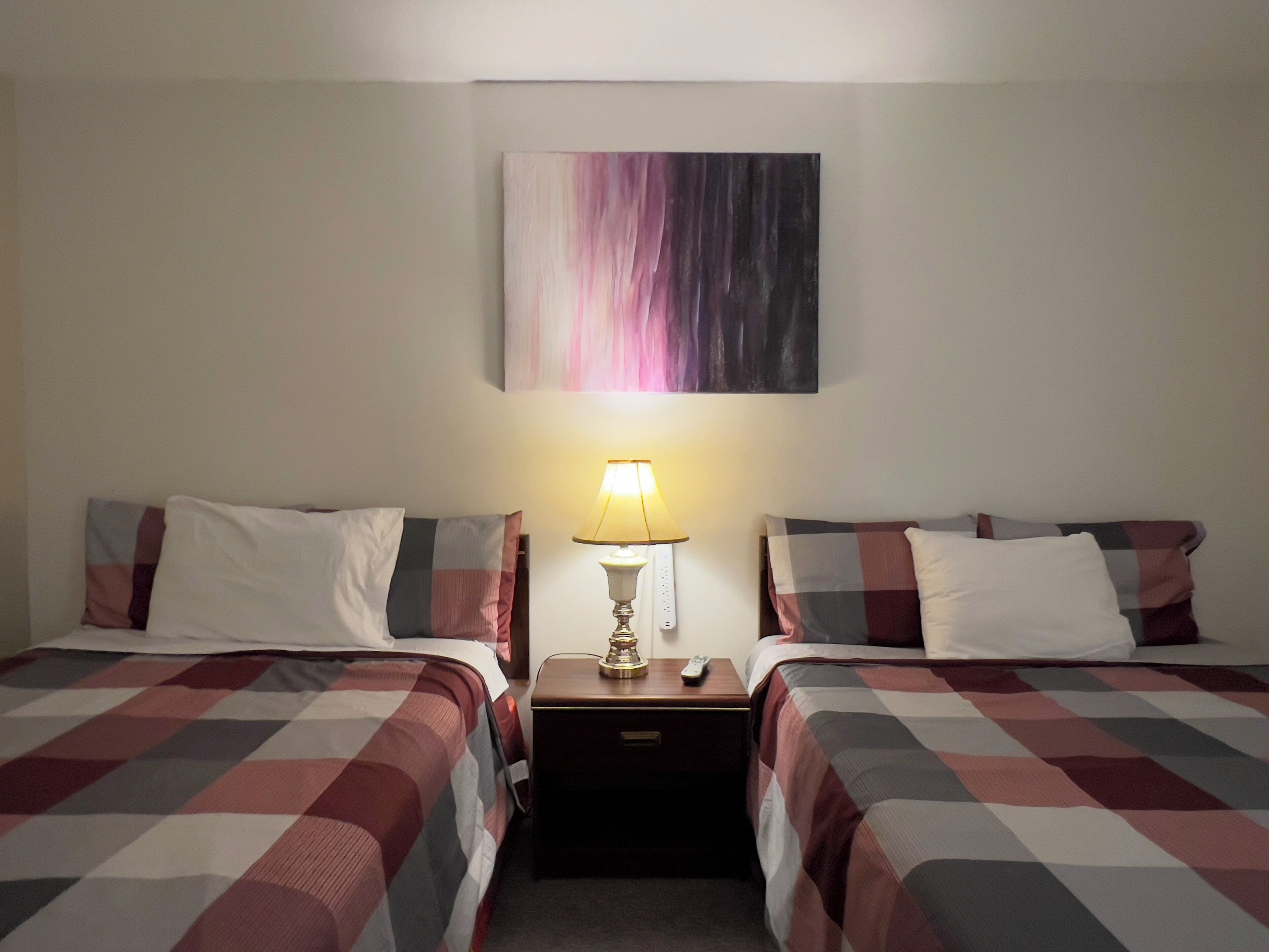 Habitación de hotel con dos camas, cada una con una colcha a cuadros, una mesita de noche con lámpara y un cuadro en la pared.