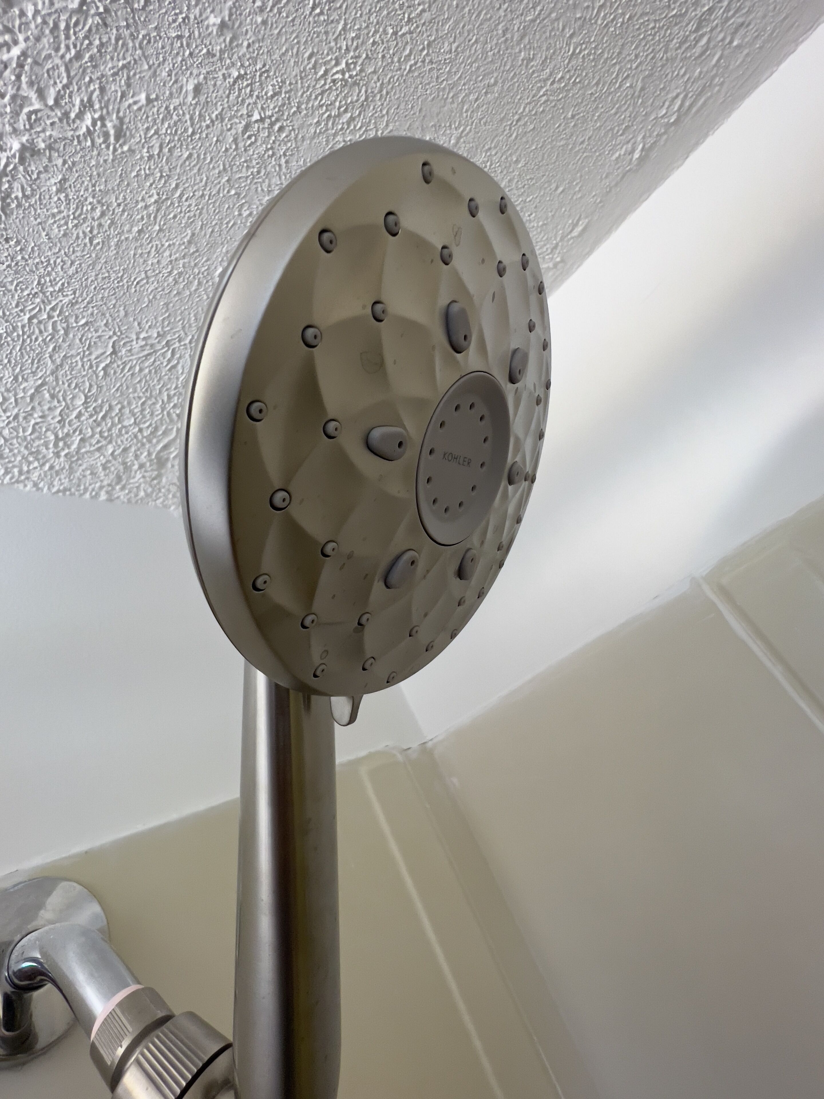 A showerhead with multiple nozzles, attached to a metal pipe, in a bathroom with white walls and a white bathtub.