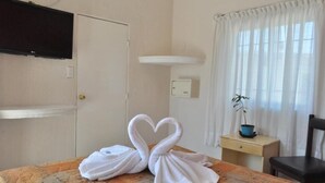 Une chambre d’hôtel avec un lit, un téléviseur fixé au mur, une table de chevet avec une plante et une fenêtre avec des rideaux.