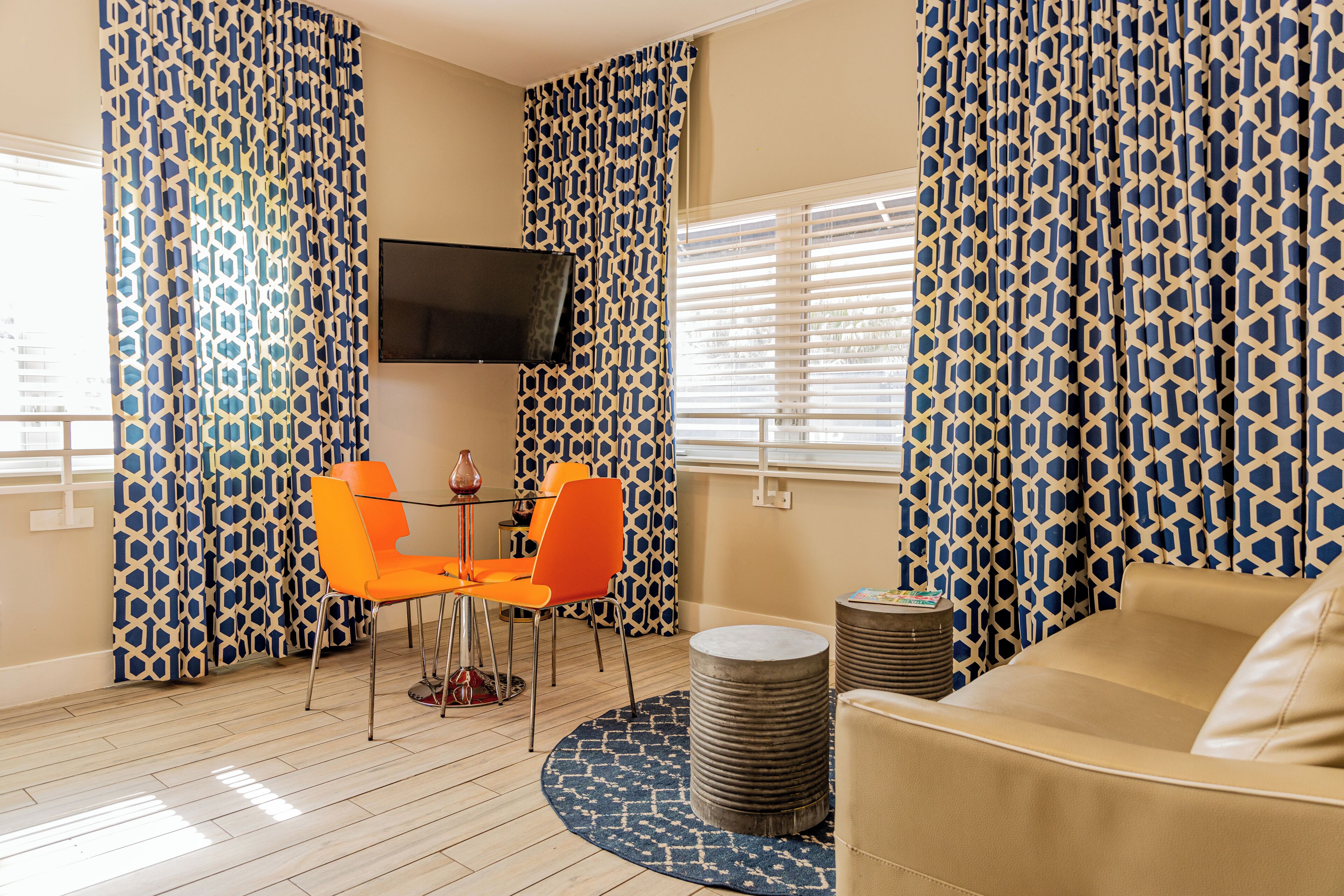 A room with a round table, two orange chairs, a beige sofa, a TV mounted on the wall, and patterned curtains.