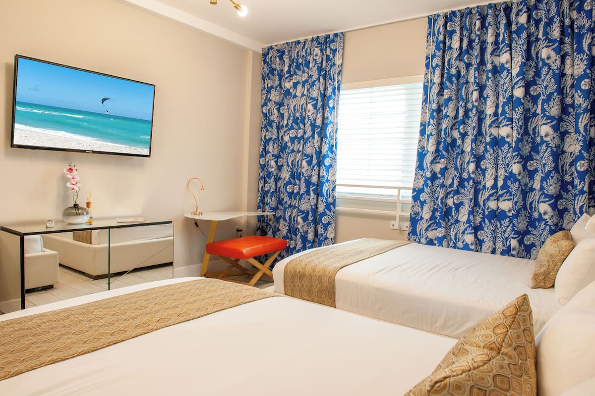 Une chambre d’hôtel avec deux lits, un bureau, une chaise, un téléviseur diffusant une scène de plage et des rideaux à motifs floraux bleus.