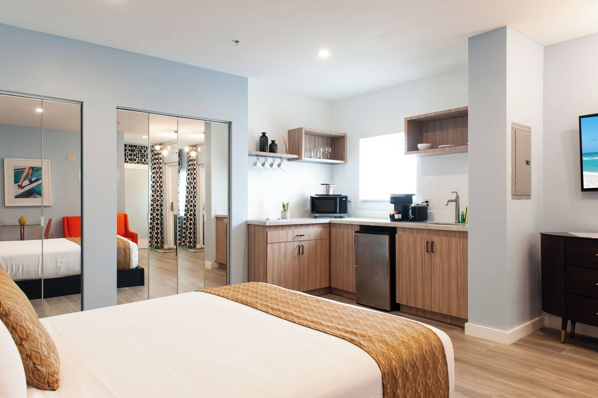 Une chambre d’hôtel moderne équipée d’un lit, d’un petit coin cuisine et d’une salle de bain visible à travers une porte en verre.