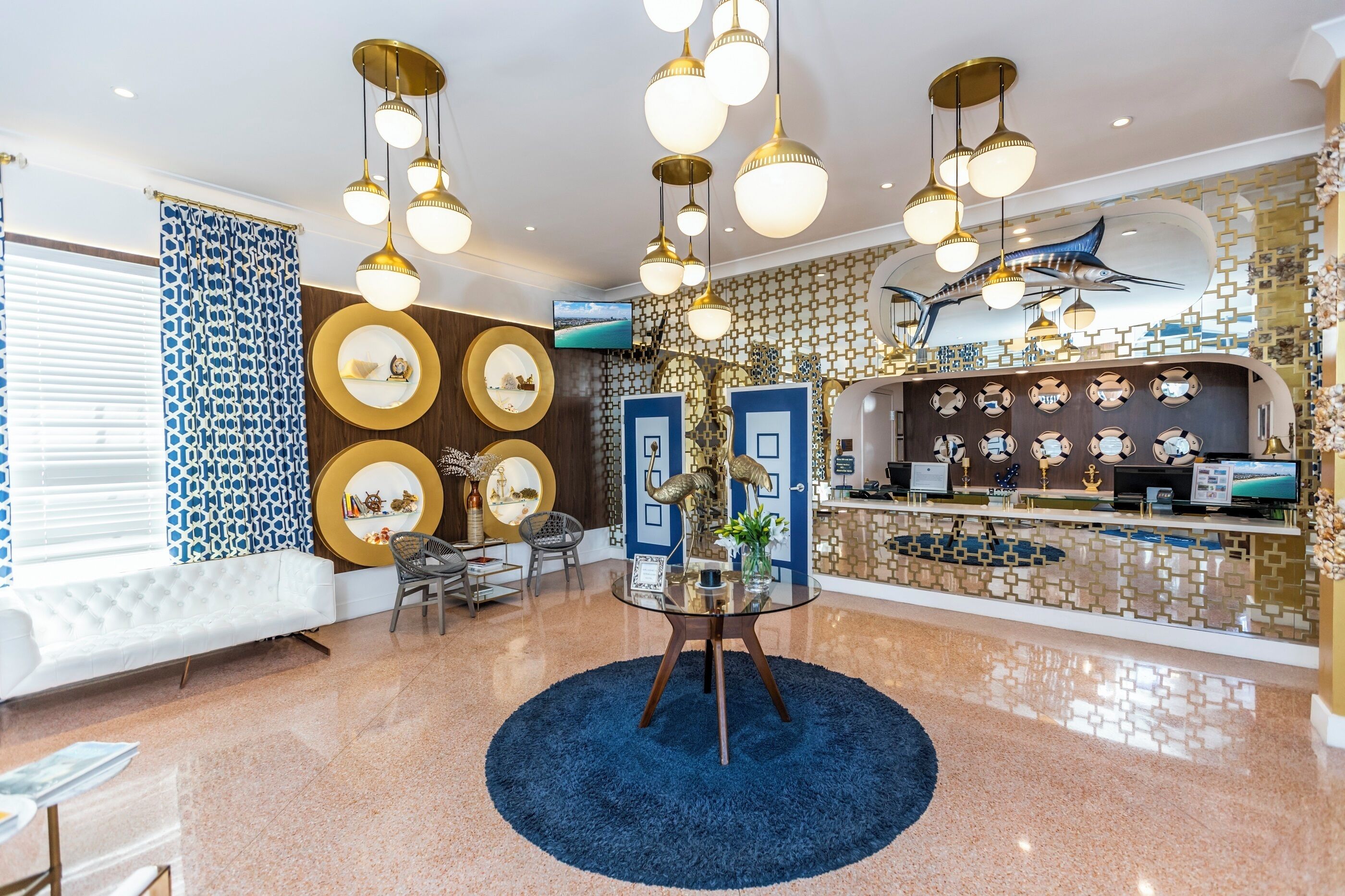 A hotel lobby with a round glass table, multiple hanging light fixtures, a reception desk with a fish mural, and a patterned wall.