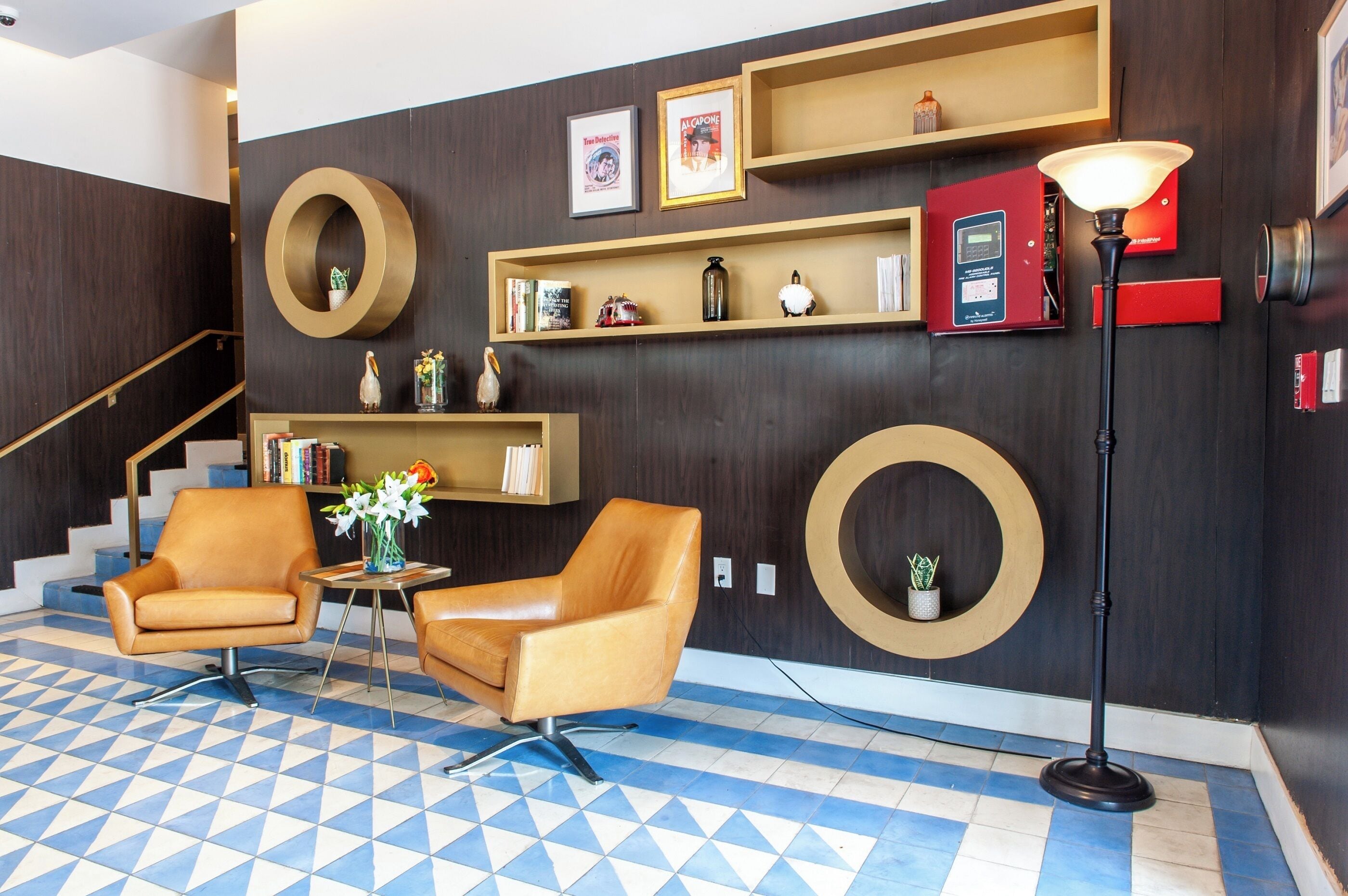 A lobby with two leather armchairs, a small table with a vase of flowers, a standing lamp, and a staircase with a dark wooden wall and blue and white patterned floor.