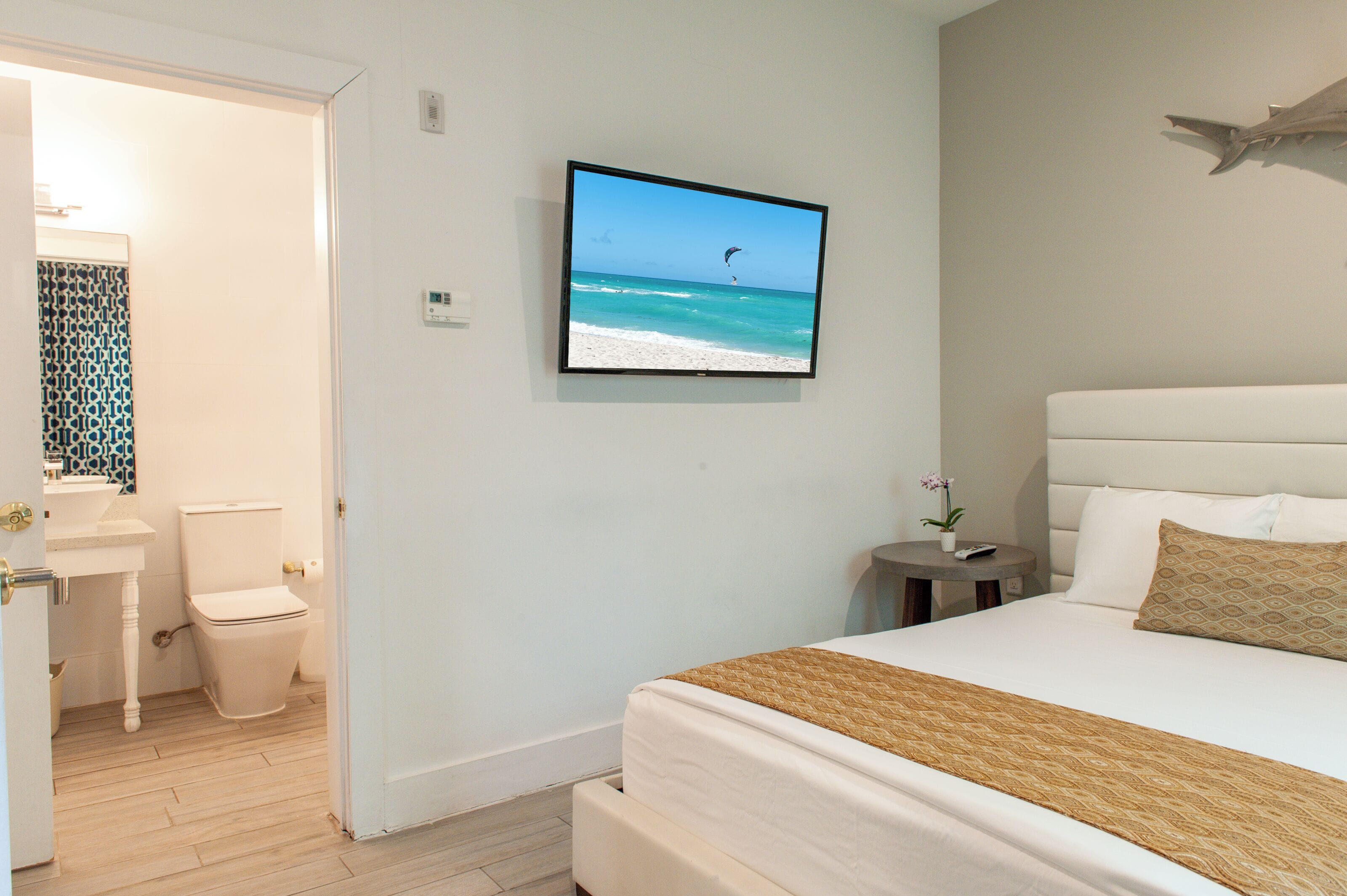 A bedroom with a bed, a television displaying a beach scene, a small table with a plant, and a bathroom with a toilet and sink.