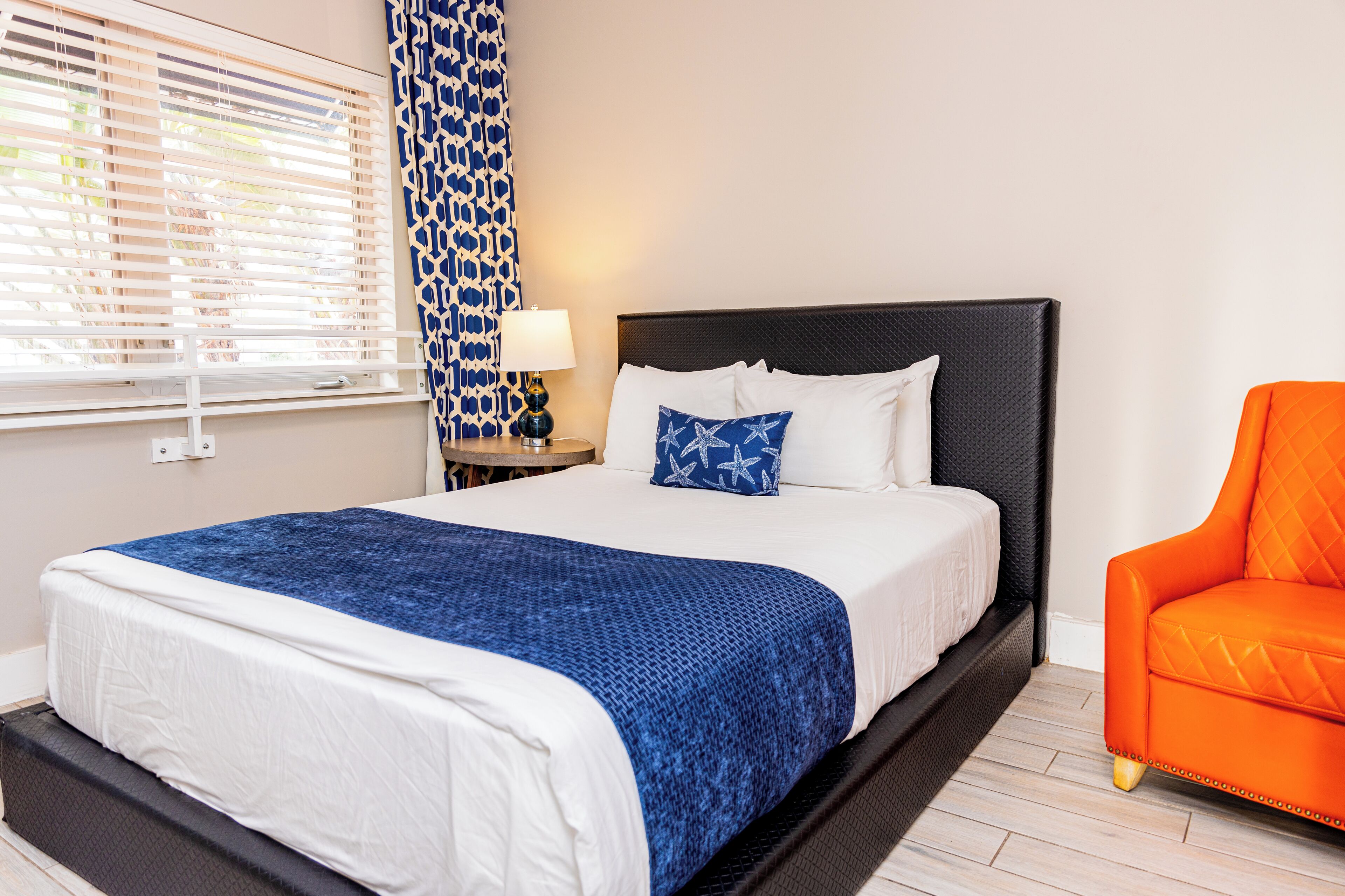 Una habitación de hotel con una cama grande, una mesita de noche con lámpara, una cortina azul estampada y un sillón naranja.