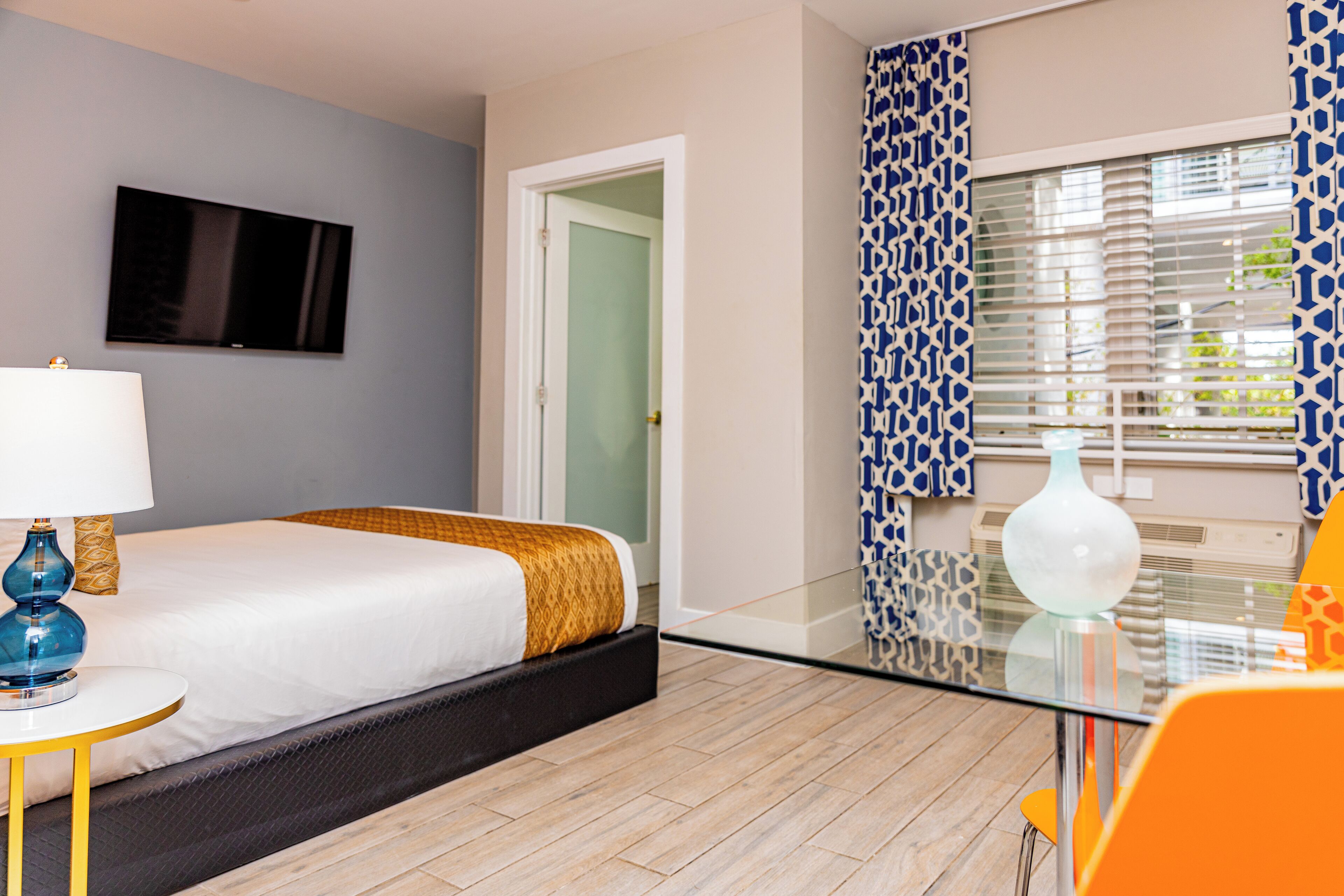 Quarto de hotel com cama, televisão na parede, mesa de vidro e janela com cortinas estampadas.