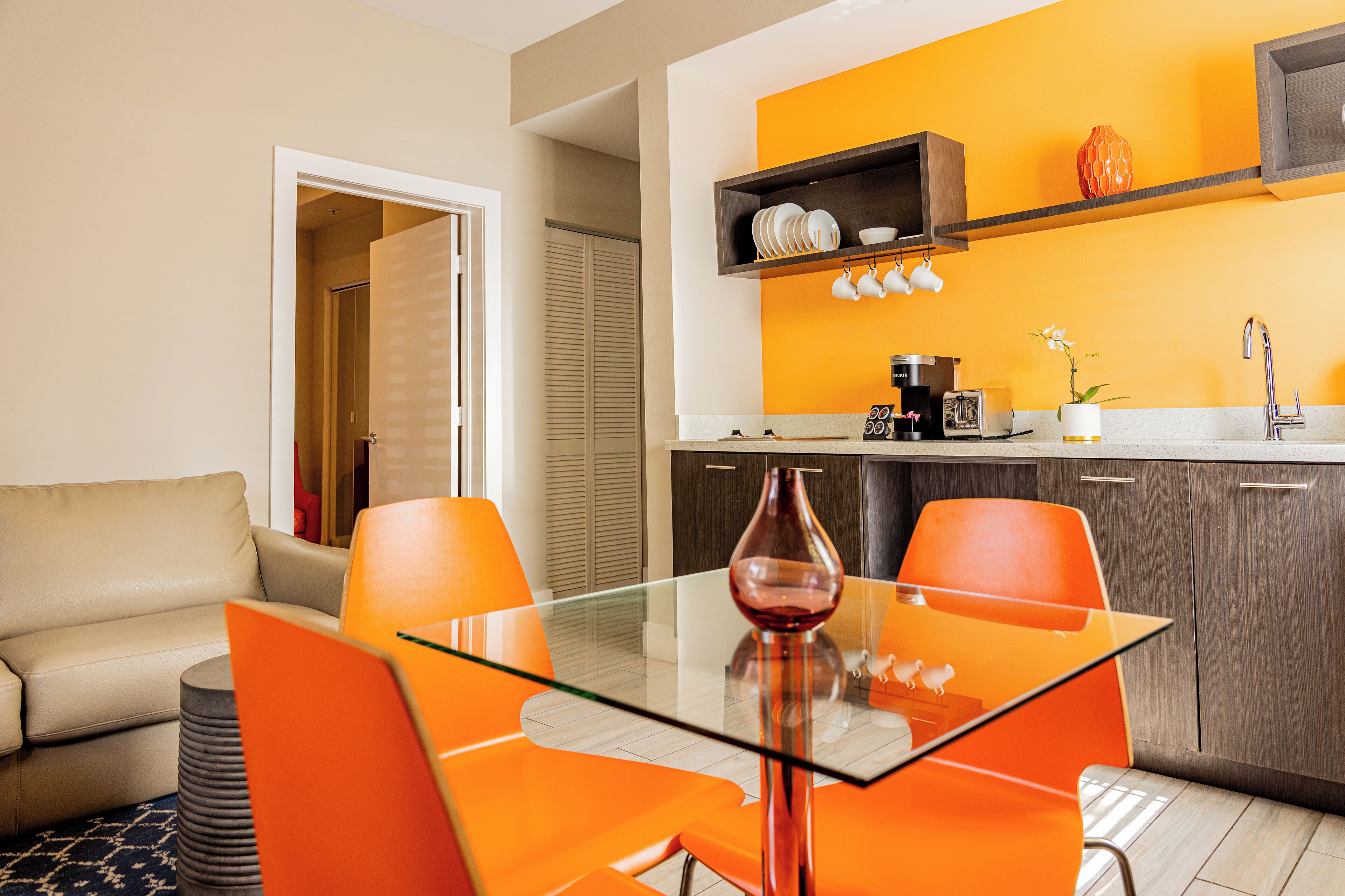 A modern kitchen with a glass table and orange chairs, a beige sofa, and a wooden cabinet with dishes.