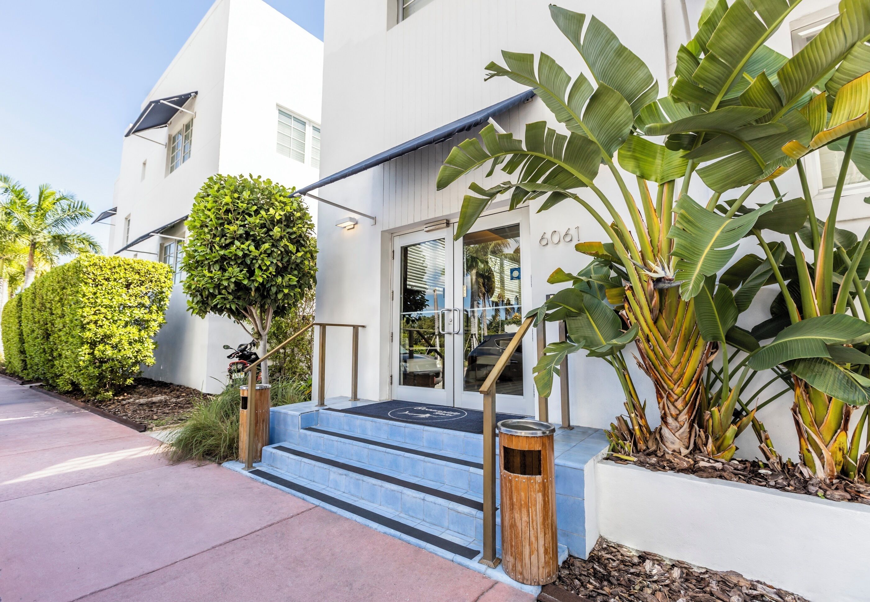 Un bâtiment moderne à la façade blanche, portant le numéro 6061, avec un escalier bleu menant à une entrée en verre, le tout entouré d’une végétation luxuriante, notamment des bananiers.