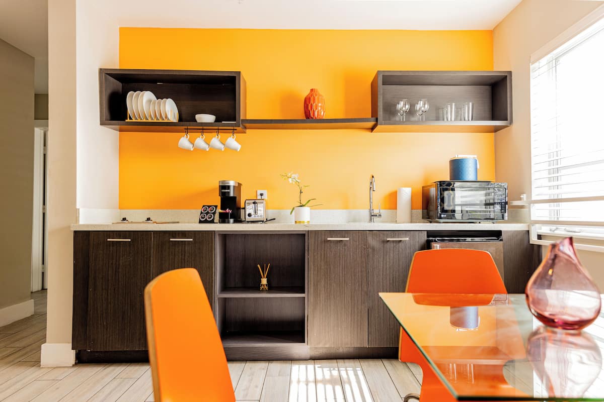 Une cuisine moderne avec des meubles en bois, un mur d’accent d’un orange vif, et une table à manger en verre accompagnée de chaises orange.