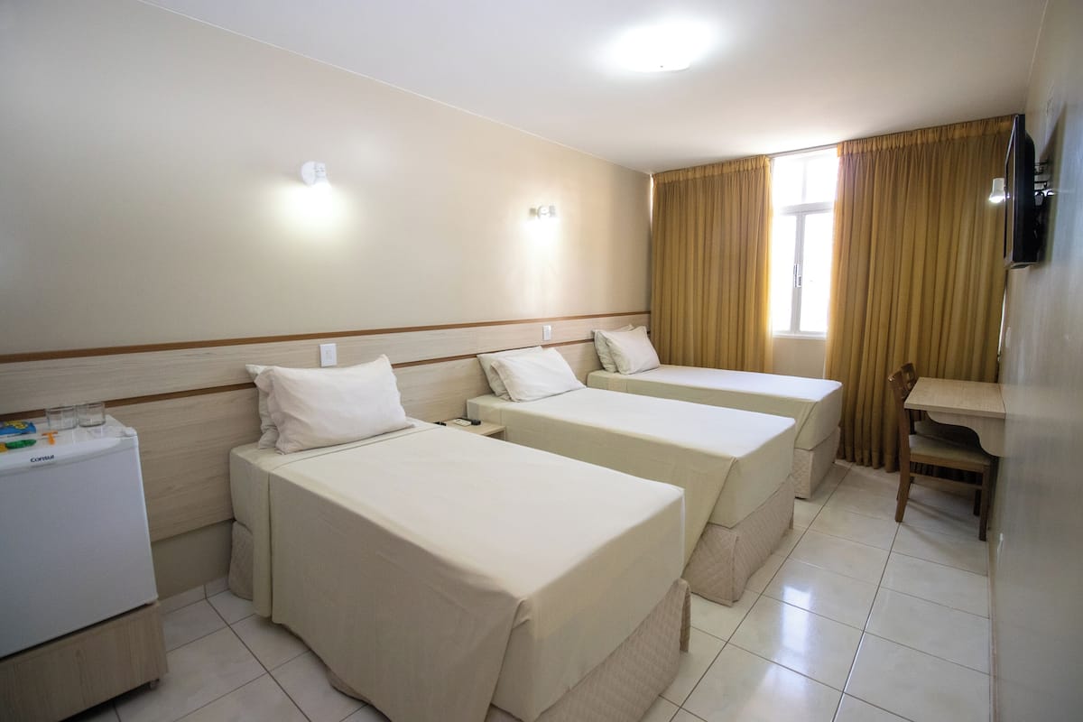 Quarto de hotel com três camas de solteiro, uma escrivaninha pequena, uma cadeira e um refrigerador.