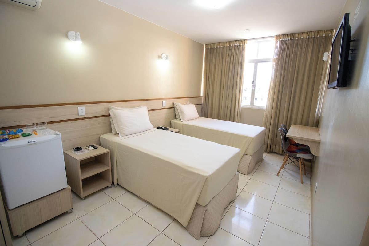 Quarto de hotel com duas camas, uma escrivaninha, uma cadeira, um frigorífico e uma televisão.
