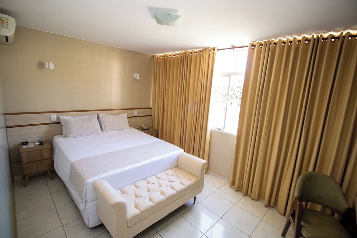 Quarto de hotel com uma cama grande, uma mesa de cabeceira, uma cadeira e uma janela com cortinas.