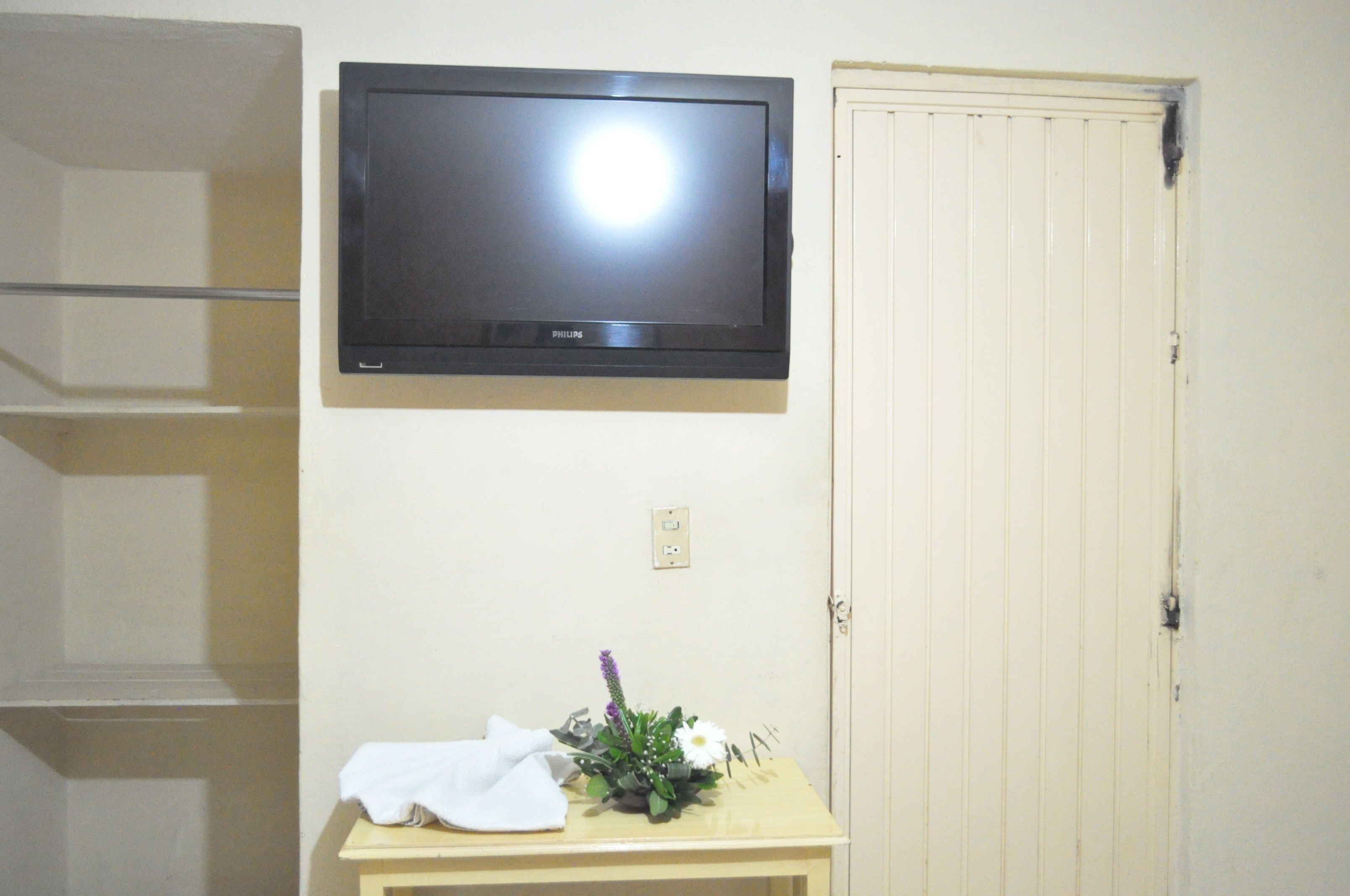 A room with a mounted television, a door, and a small table with a white cloth and a floral arrangement.