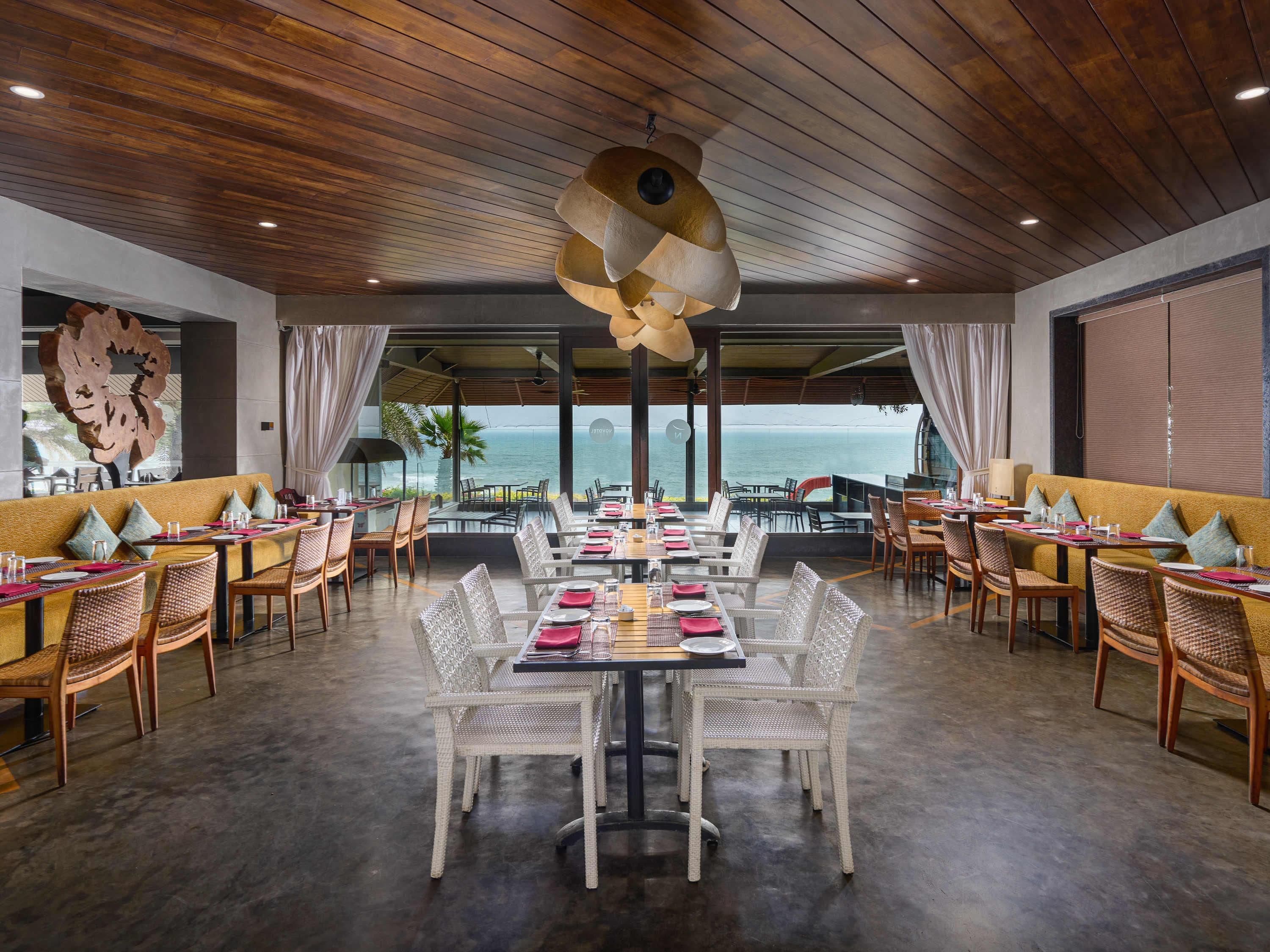 A restaurant with a view of the ocean, featuring wooden ceiling, white wicker chairs, and tables set with pink napkins.
