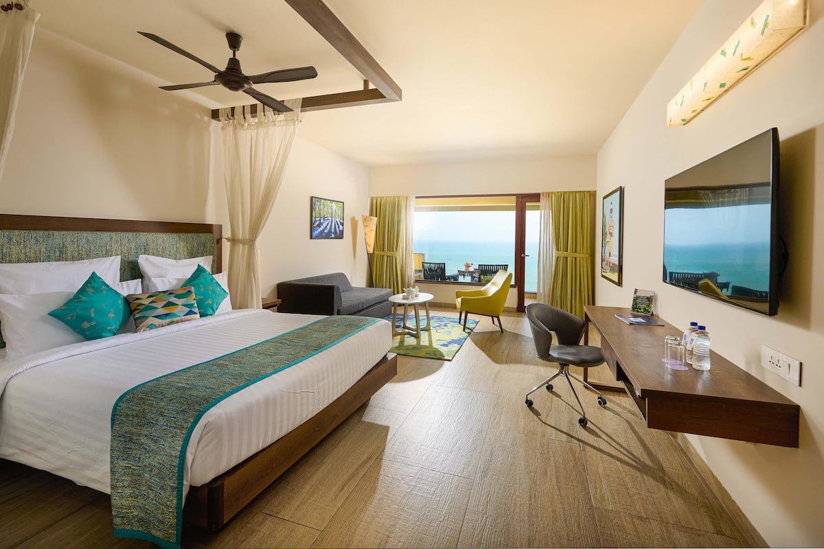 Ein Hotelzimmer mit einem großen Bett, einem Schreibtisch, einem Stuhl, einem Fernseher und einem Balkon mit Meerblick.
