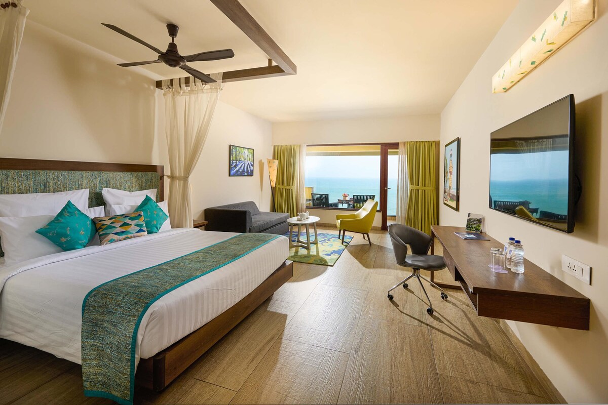A hotel room with a large bed, a desk, a chair, a TV, and a balcony with a view of the sea.