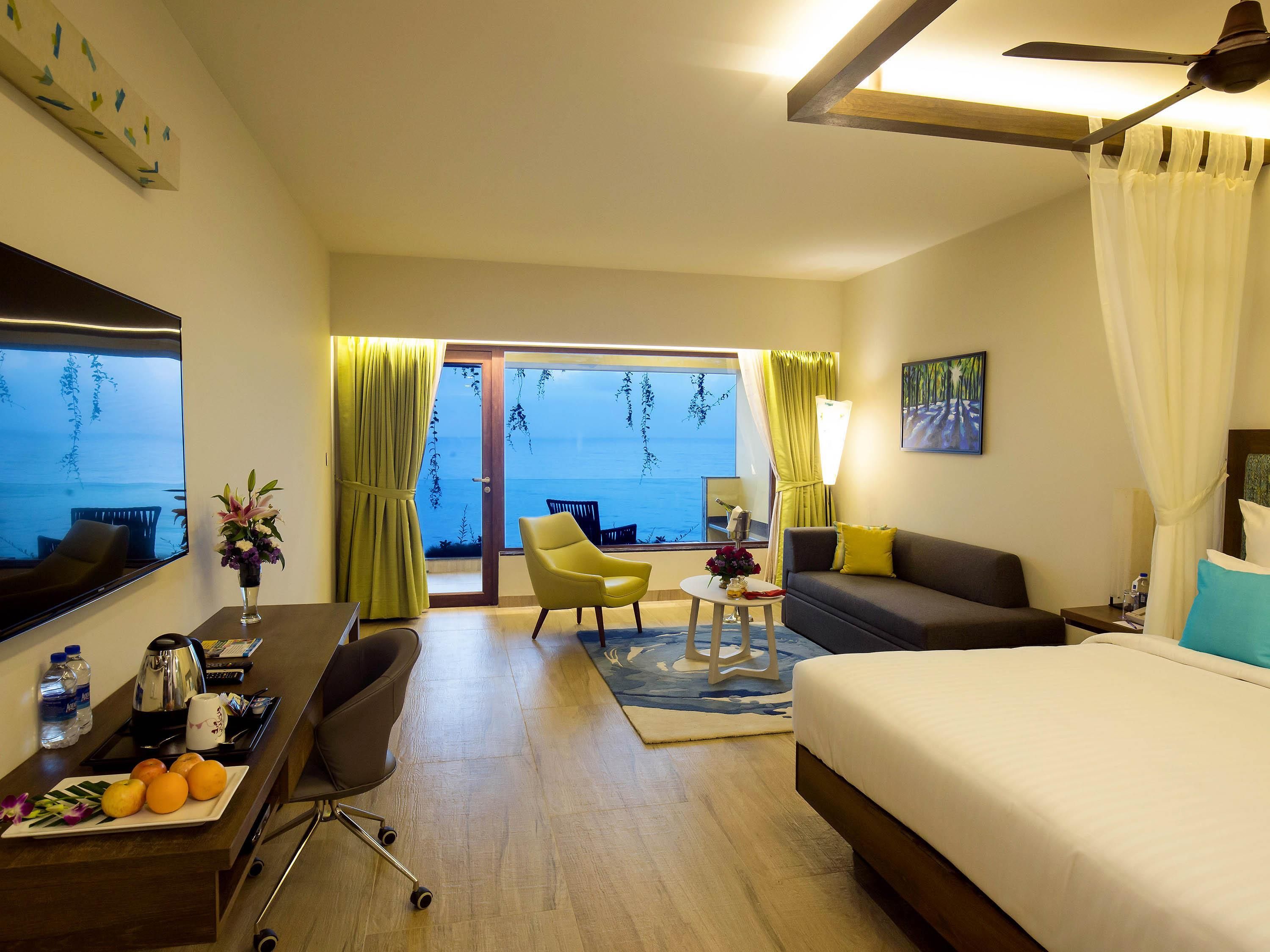A hotel room with a bed, a desk with a chair, a small table with a lamp, a sofa, and a view of the sea through large windows.