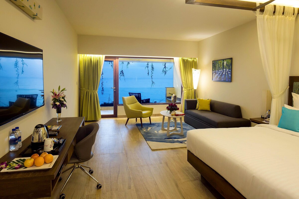 A hotel room with a bed, a desk with a chair, a small table with a lamp, a sofa, and a view of the sea through large windows.