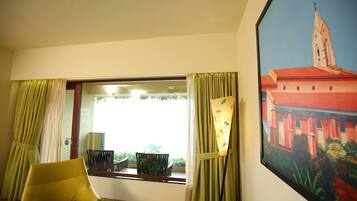 A room with a large window, a yellow armchair, and a framed painting of a building.
