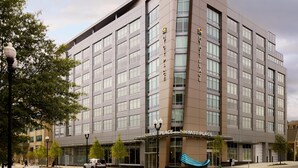 Un hôtel à plusieurs étages au design moderne, doté d’une entrée imposante avec un auvent bleu et d’un grand logo.