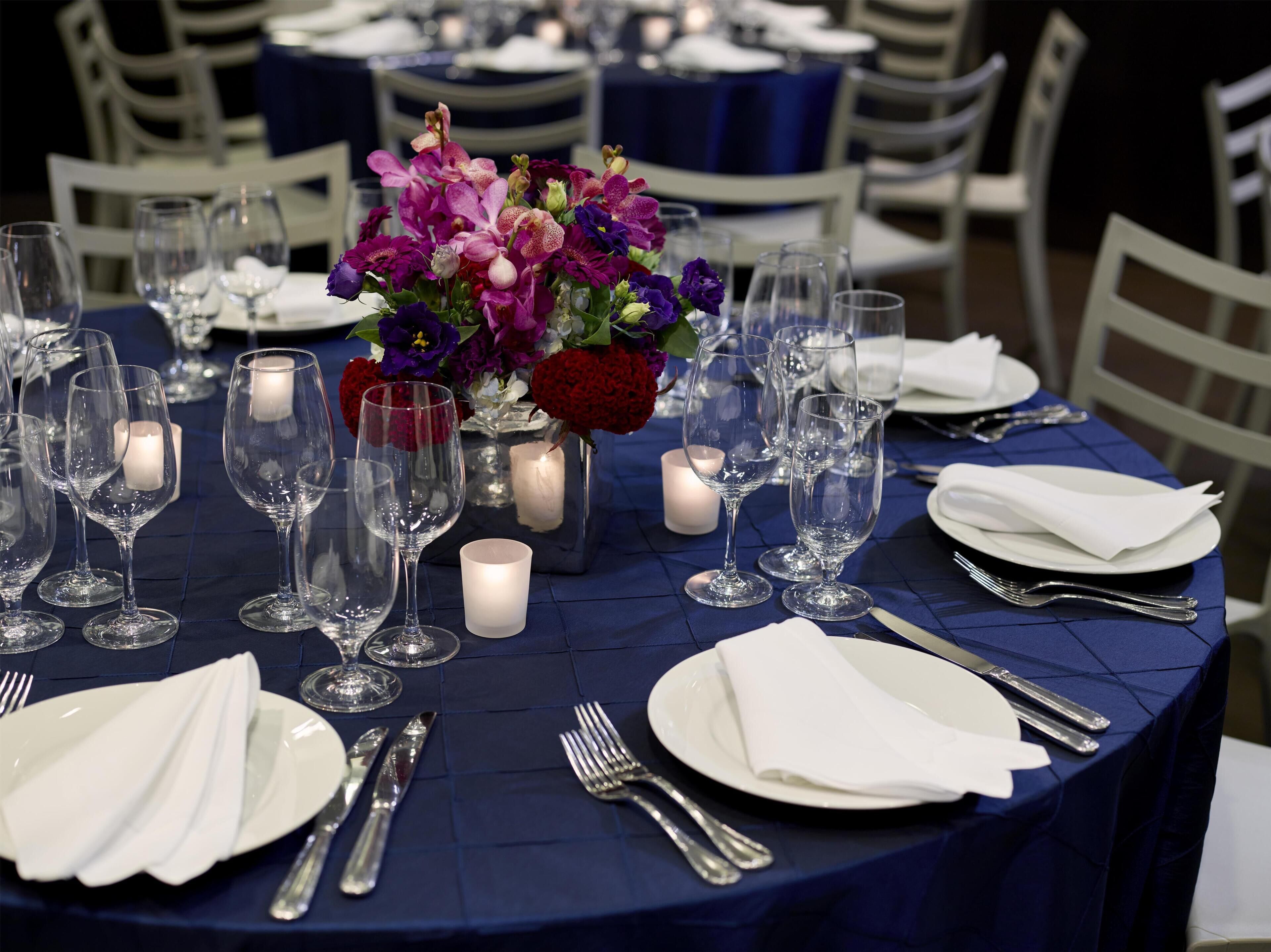 A table set with white plates, silverware, and clear wine glasses, surrounded by candles and a vase of purple flowers.