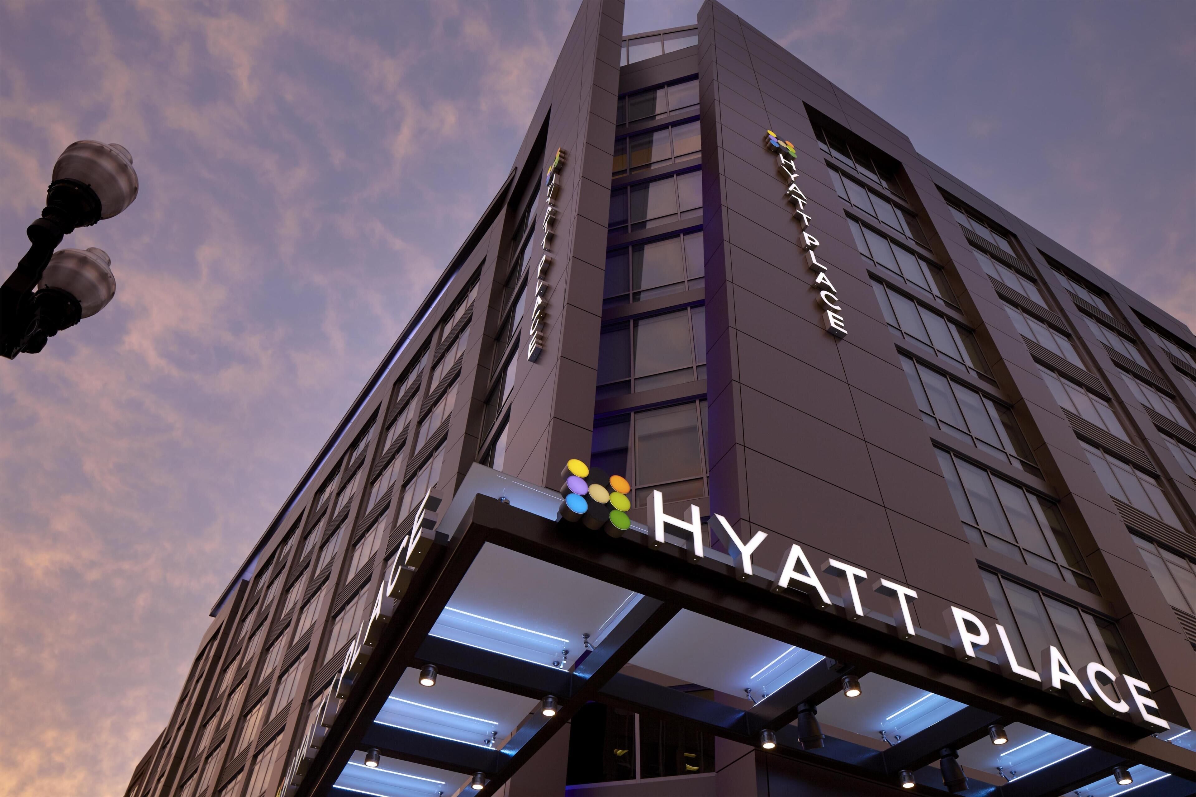 Um edifício moderno com a placa 'HYATT PLACE', destacando uma entrada imponente com fachada de vidro e uma fileira de luzes acima.