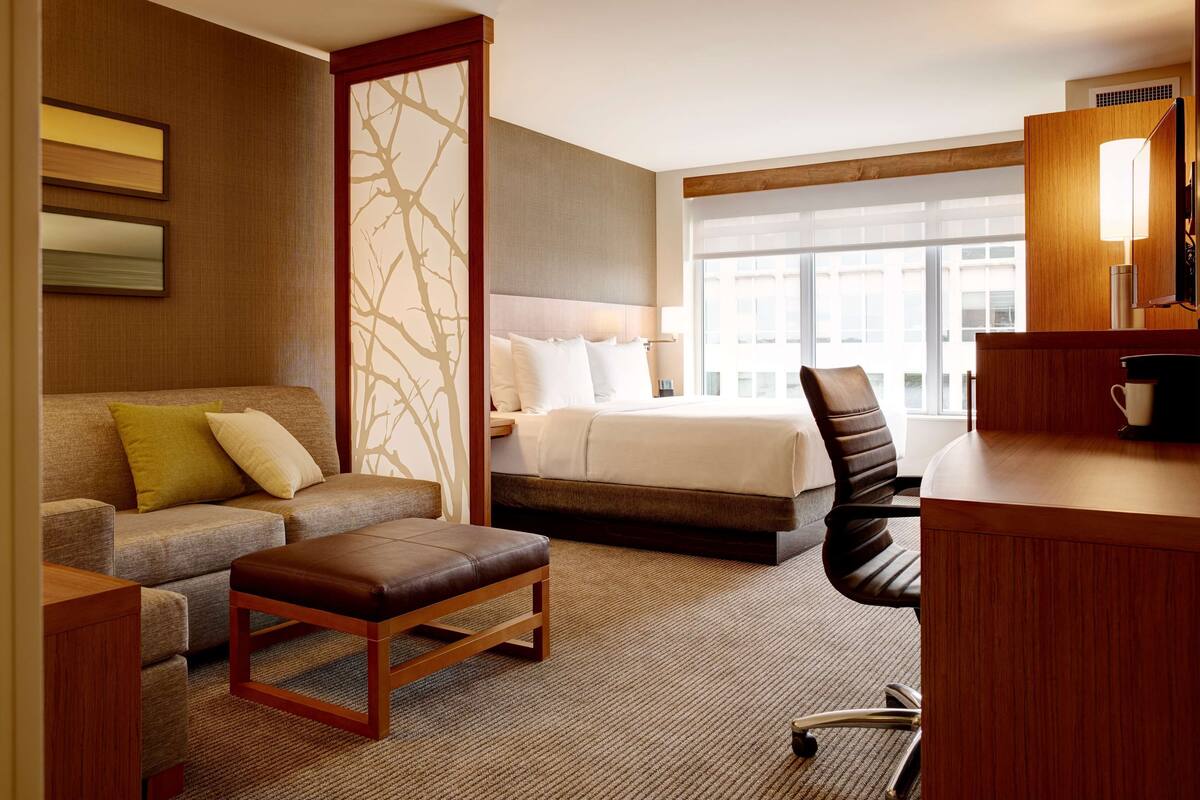 A hotel room with a bed, a sofa, a desk, and a chair. There is a large window providing a view of the city.