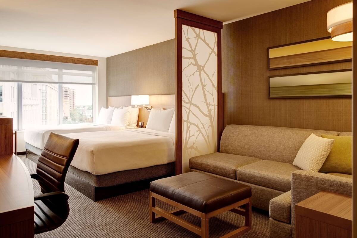 A hotel room with two beds, a sofa, an ottoman, a desk, and a large window offering a city view.