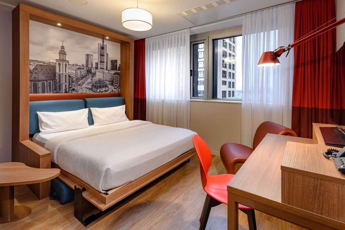 A hotel room with a bed, a desk, a chair, a small table, and a window with curtains.
