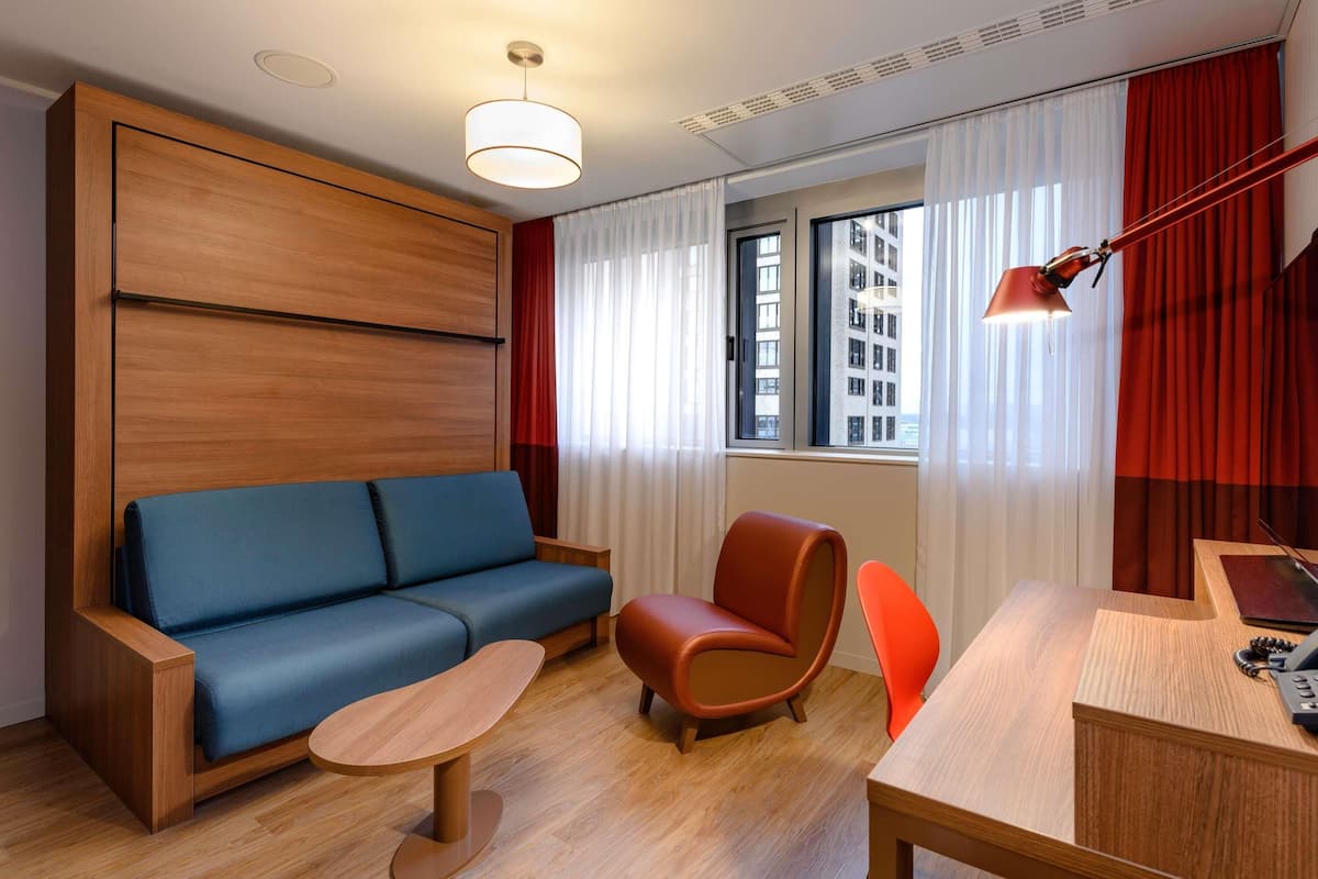 A modern hotel room with a wooden accent wall, a blue sofa, a wooden coffee table, a desk with a telephone, and a view of buildings through the window.