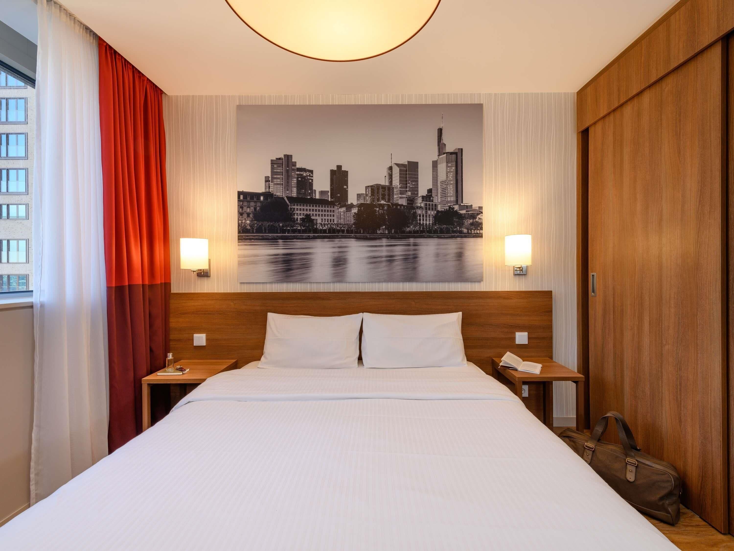 A hotel room with a bed, two bedside tables, a cityscape painting on the wall, and a view of buildings outside the window.