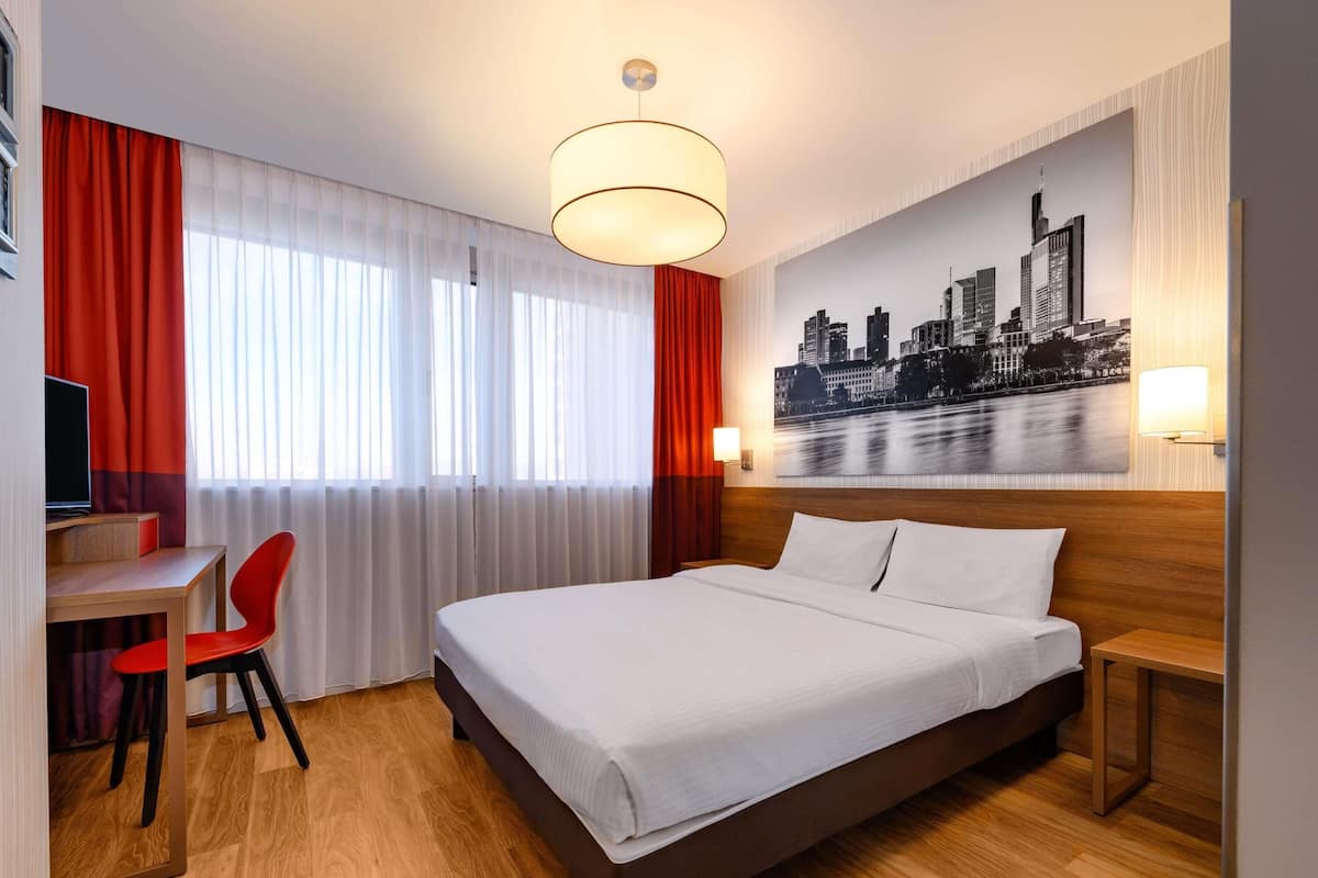 A hotel room with a bed, a desk with a chair, a cityscape painting, and a window with curtains.