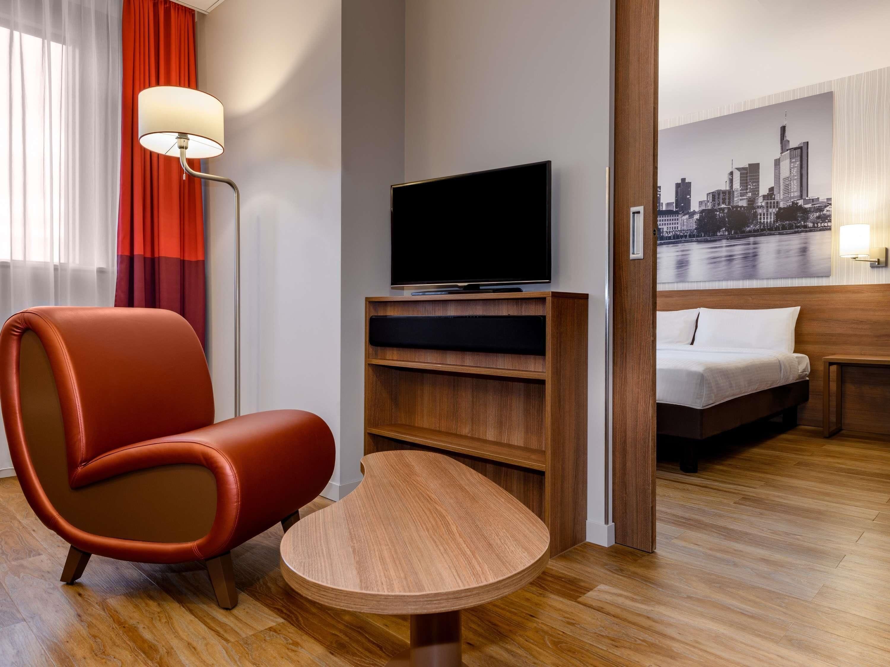 Een hotelkamer met een houten vloer, een rode lederen fauteuil, een televisie op een houten dressoir, een bed met witte lakens en een stadsgezichtschilderij aan de muur.