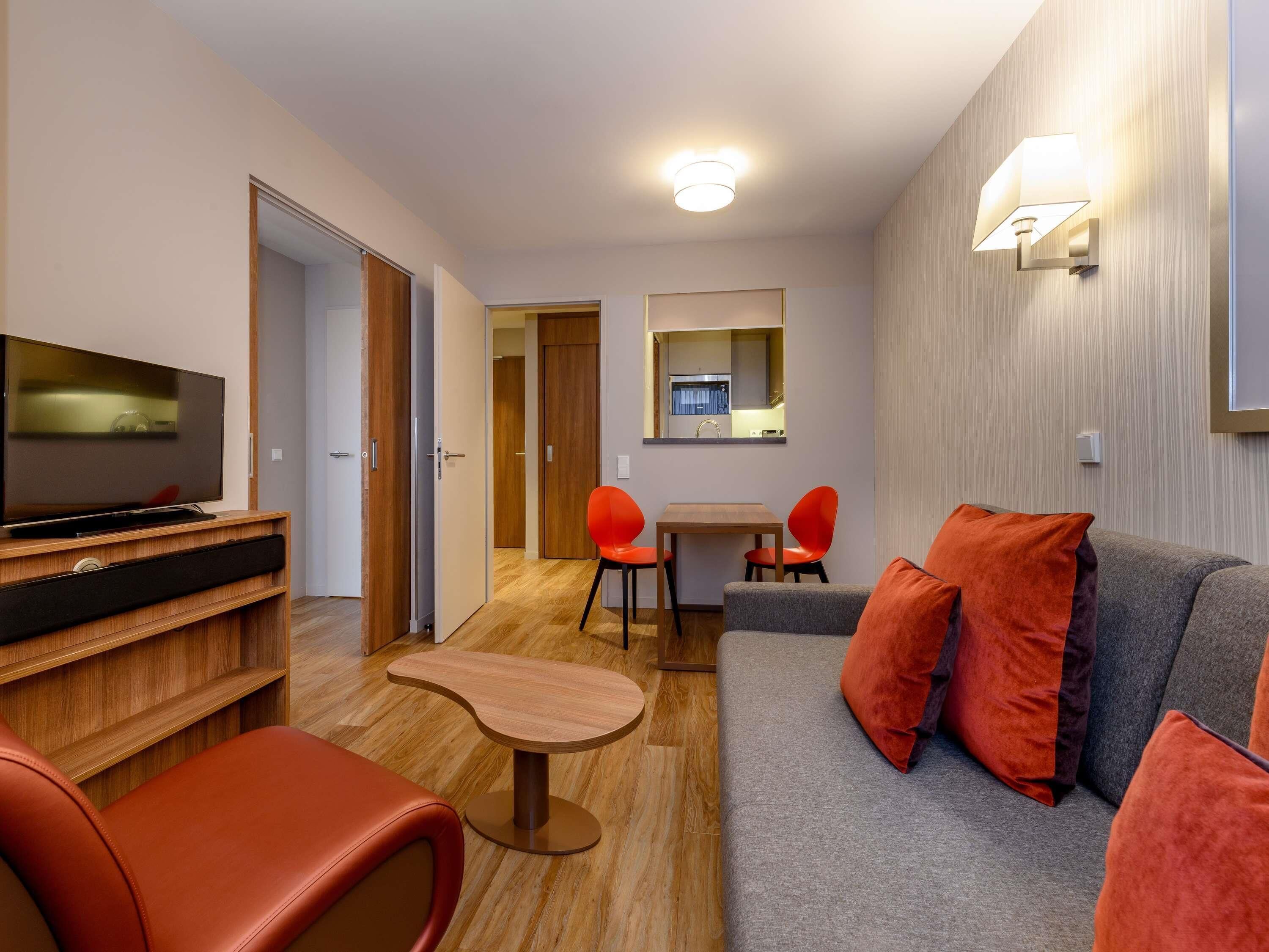Una habitación de hotel moderna con un televisor de pantalla plana, un sofá con cojines naranjas, una pequeña mesa de madera y una zona de comedor con sillas rojas.