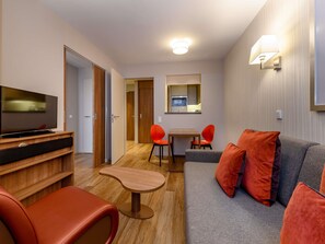 A modern hotel room with a flat-screen TV, a sofa with orange cushions, a small wooden table, and a dining area with red chairs.