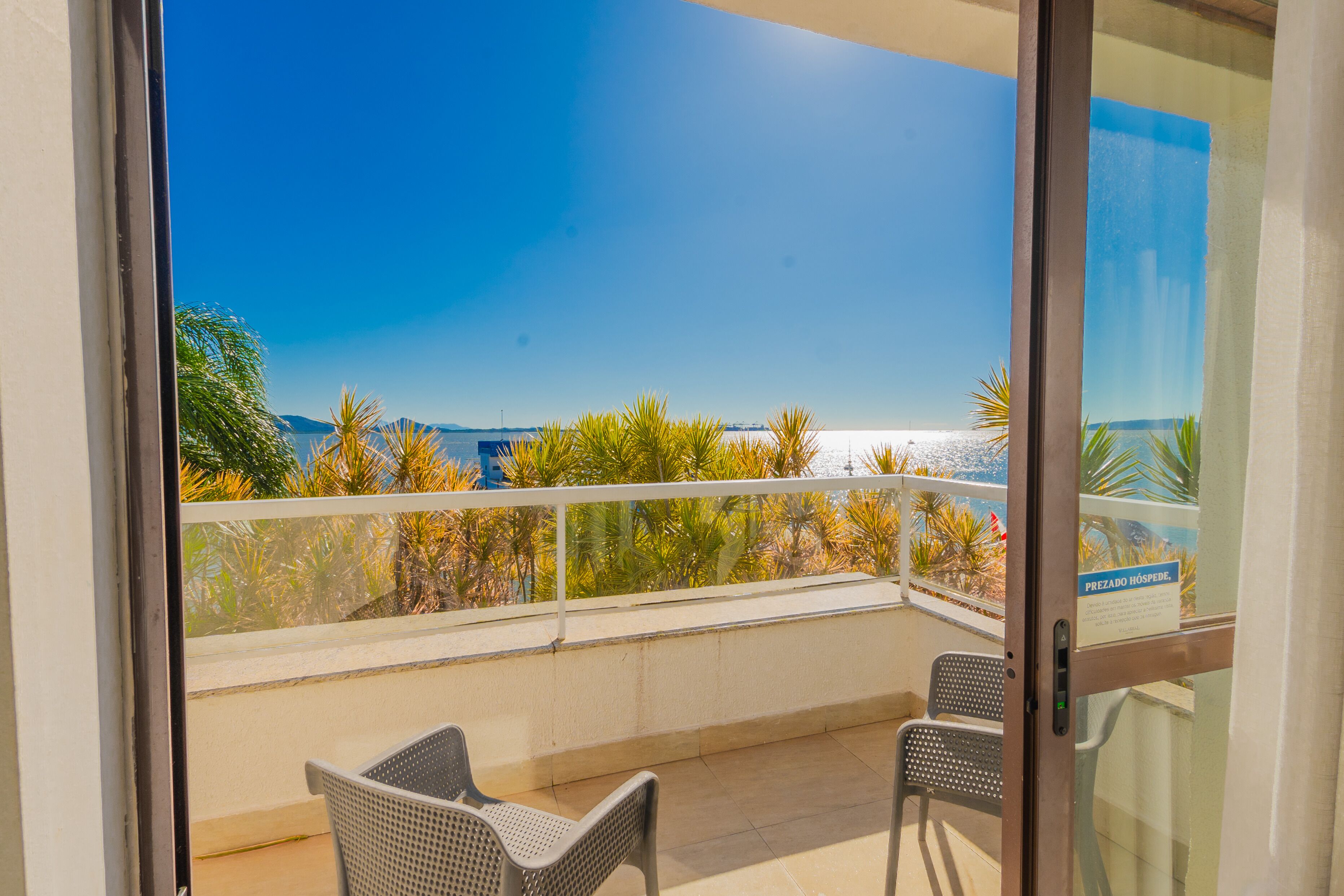 A balcony with two wicker chairs, a glass door, and a view of a beach and palm trees.