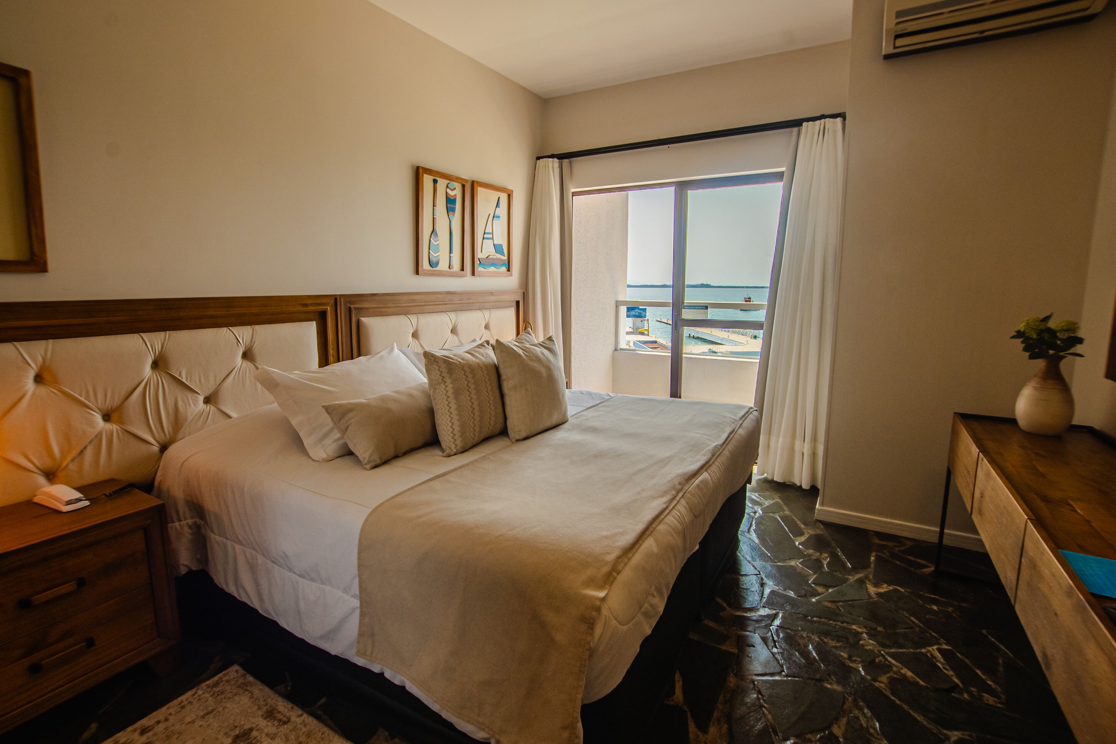 Habitación de hotel con una cama grande, una mesita de noche de madera, un escritorio y vista a la playa a través de una puerta corrediza de vidrio.