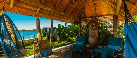 A covered outdoor seating area with wicker chairs and a hanging chair, overlooking a lush garden and a body of water.