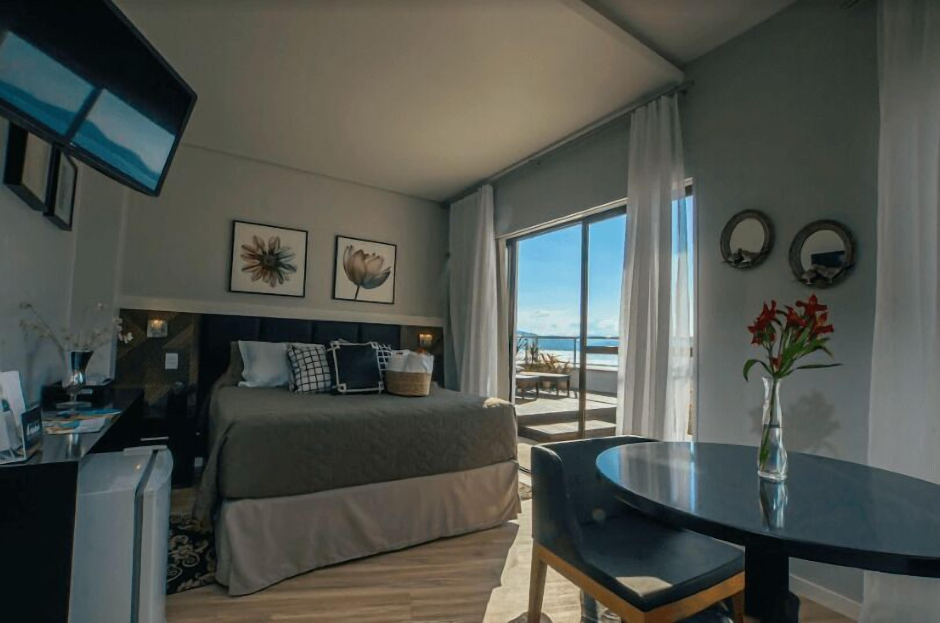 A hotel room with a bed, a desk, a chair, a TV, and a view of the beach.