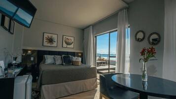 A hotel room with a bed, a desk, a chair, a TV, and a view of the beach.