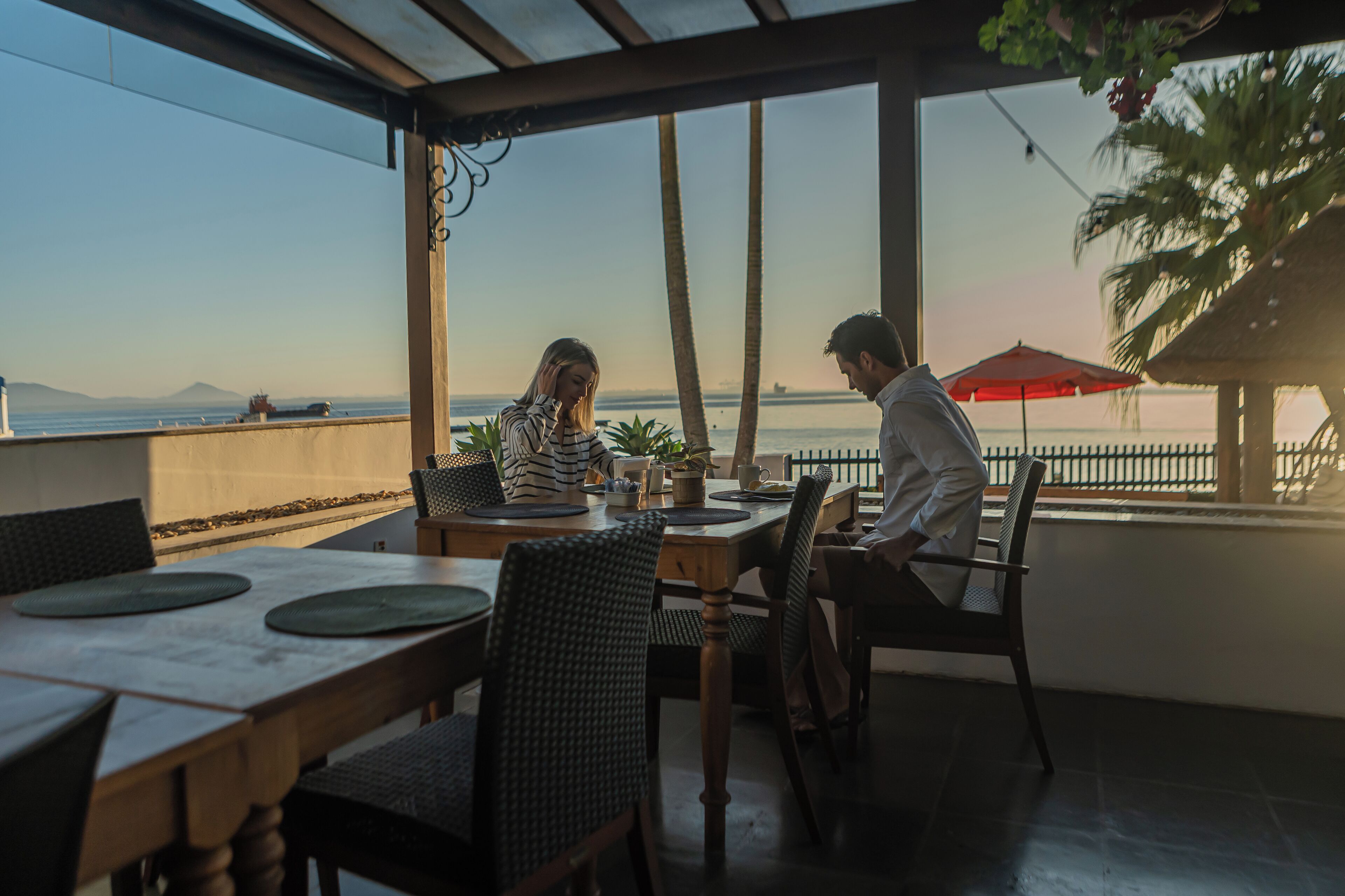A dining area with a view of the sea, featuring wooden tables and chairs, and a red umbrella.