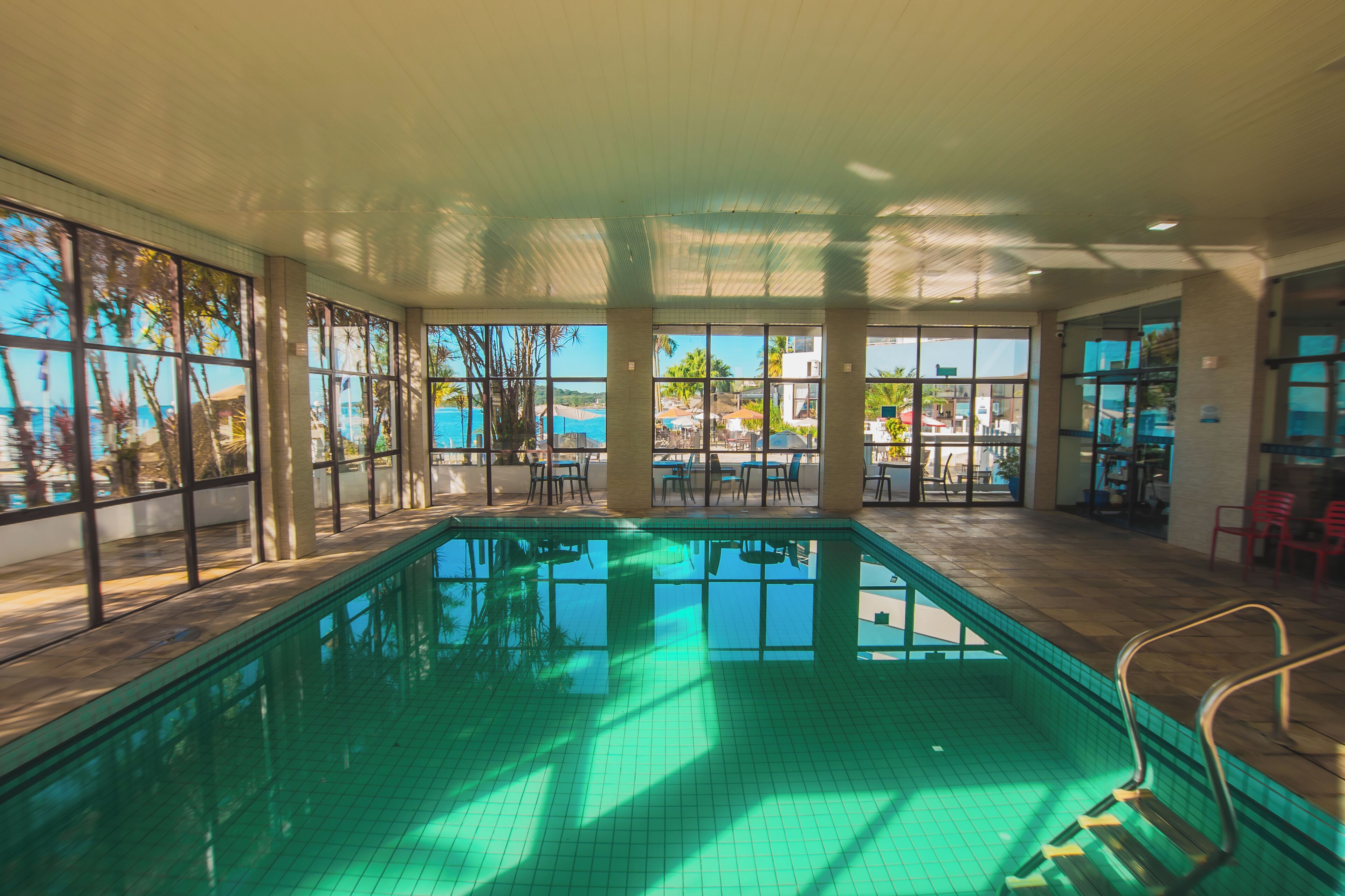 Une piscine intérieure offrant une vue dégagée sur l’océan et les palmiers grâce à de grandes fenêtres.