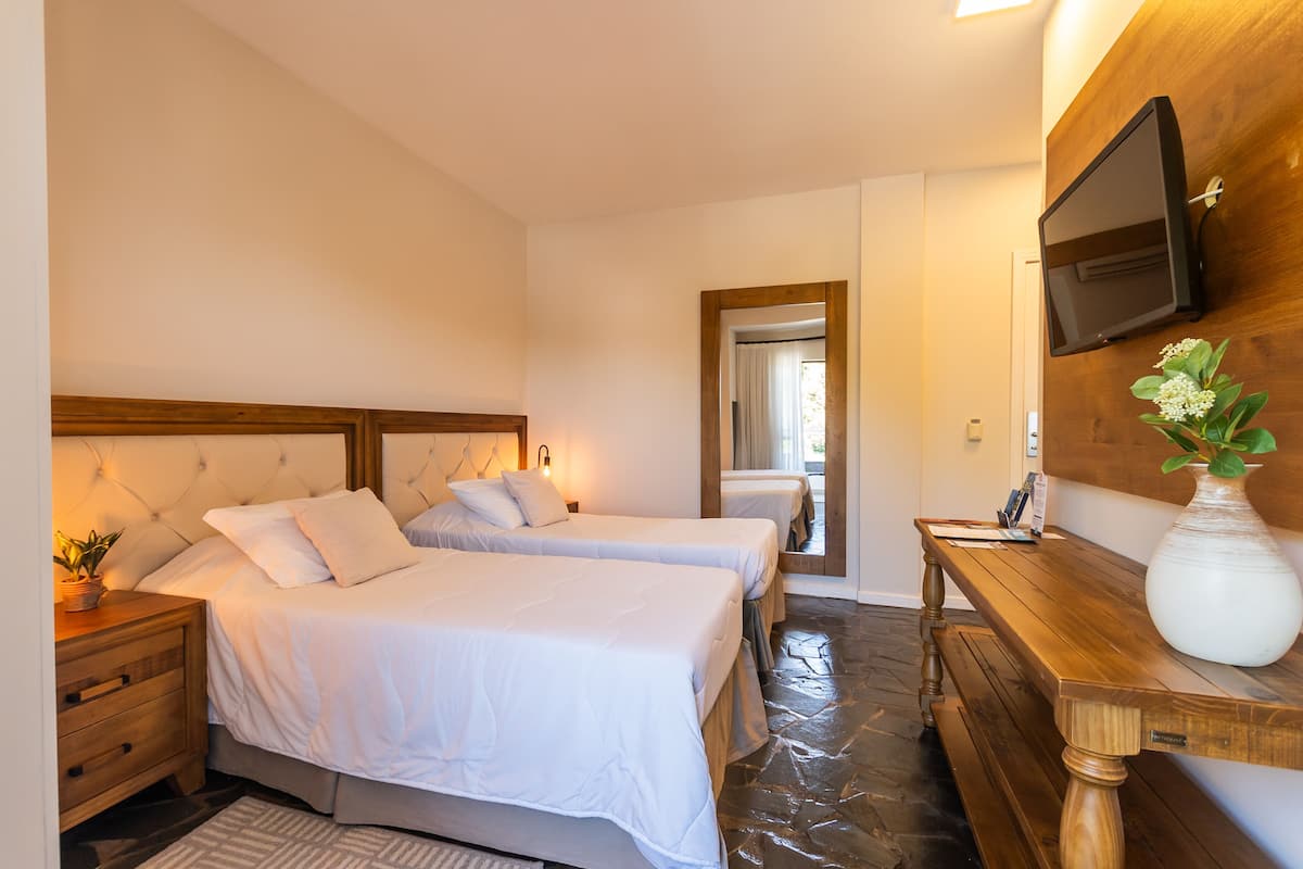 A hotel room with two beds, a wooden bench, a TV mounted on the wall, and a vase with flowers.