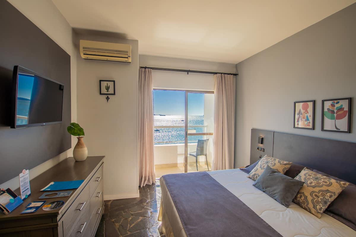 A hotel room with a bed, a desk, a TV, a plant, a balcony with a view, and artwork on the wall.